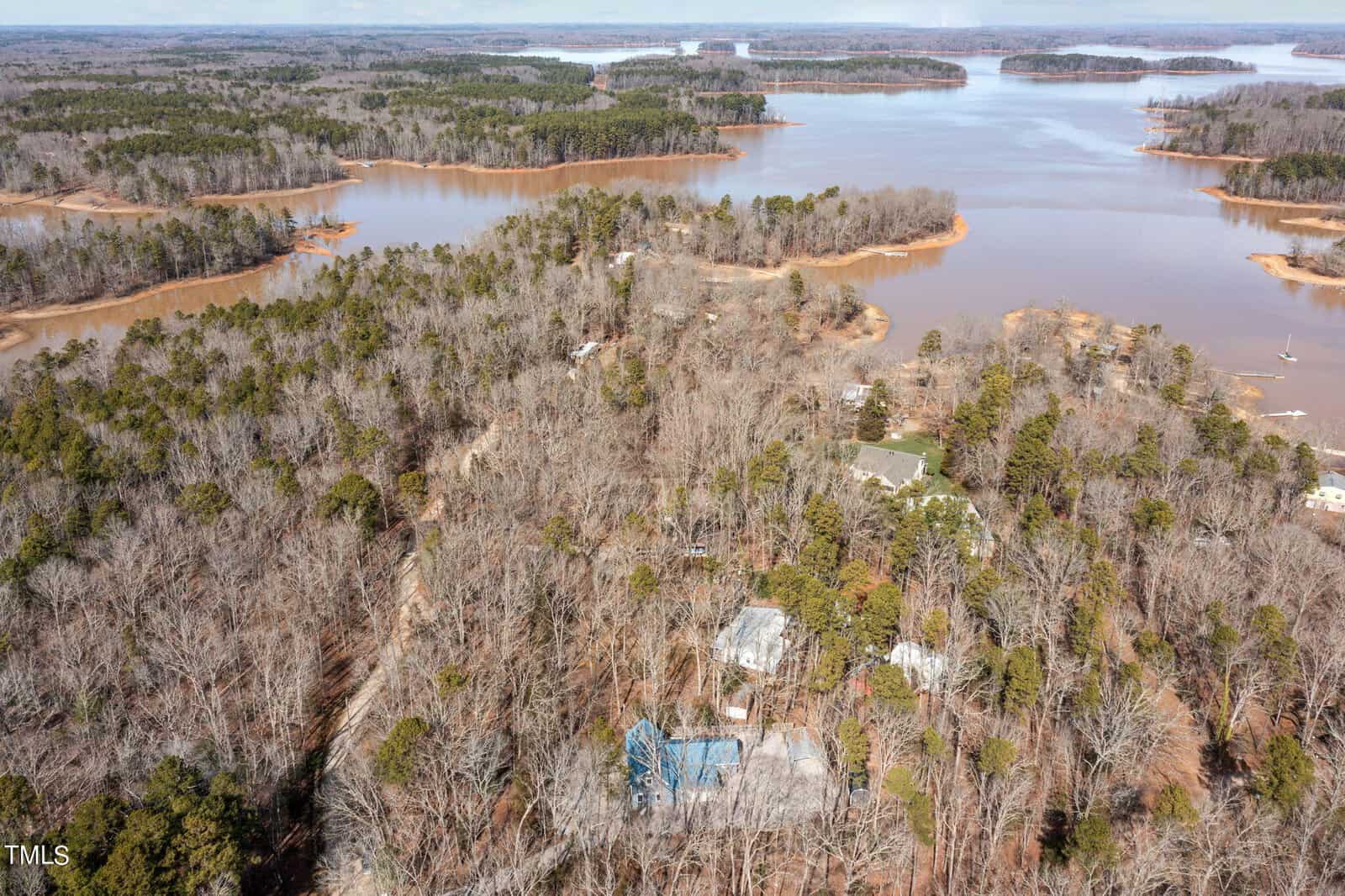 Aerial view of a wooded area with a lake and several small islands. Houses with gray and blue roofs are visible among the trees. The landscape is a mix of barren and evergreen trees near the water's edge.