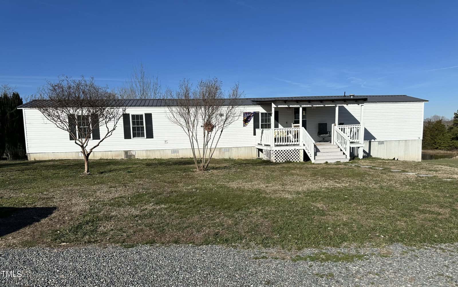 A white mobile home with a small porch and steps leading to the entrance is set in a grassy area. Two leafless trees stand in front, and there's a gravel path in the foreground beneath a clear blue sky.