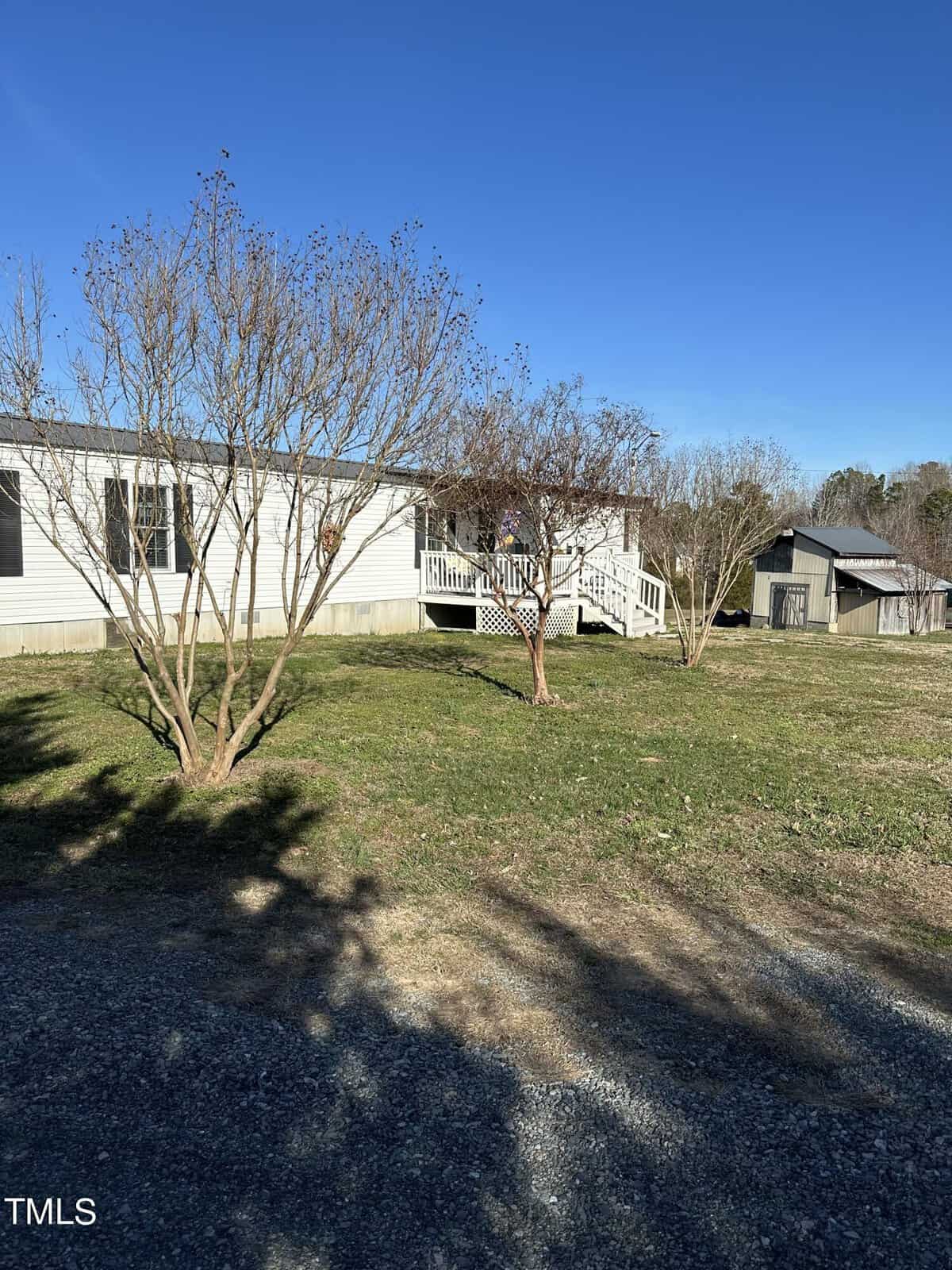 A white manufactured home with a porch is surrounded by a grassy yard and bare trees. A gravel driveway is in the foreground, and a small shed is in the background under a clear blue sky.