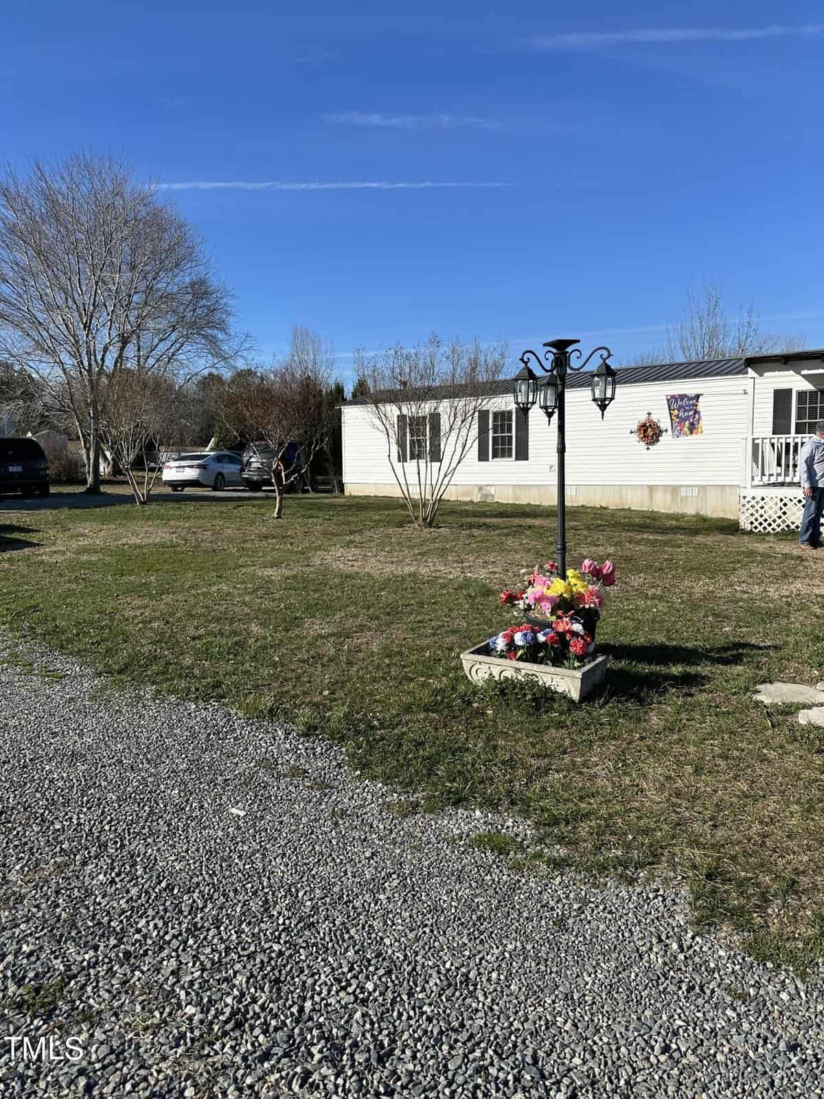 A white mobile home with a porch sits on a green lawn. A lamp post with hanging flower pots is in the foreground. Cars are parked nearby, and leafless trees frame the scene under a clear blue sky.