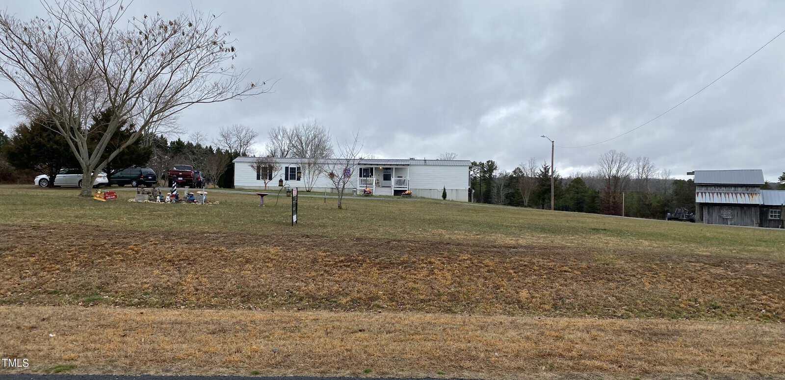 A white mobile home with a small porch sits atop a grassy hill. Several cars are parked nearby, and a tree without leaves is in the foreground. The sky is overcast, adding a gray tone to the scene.