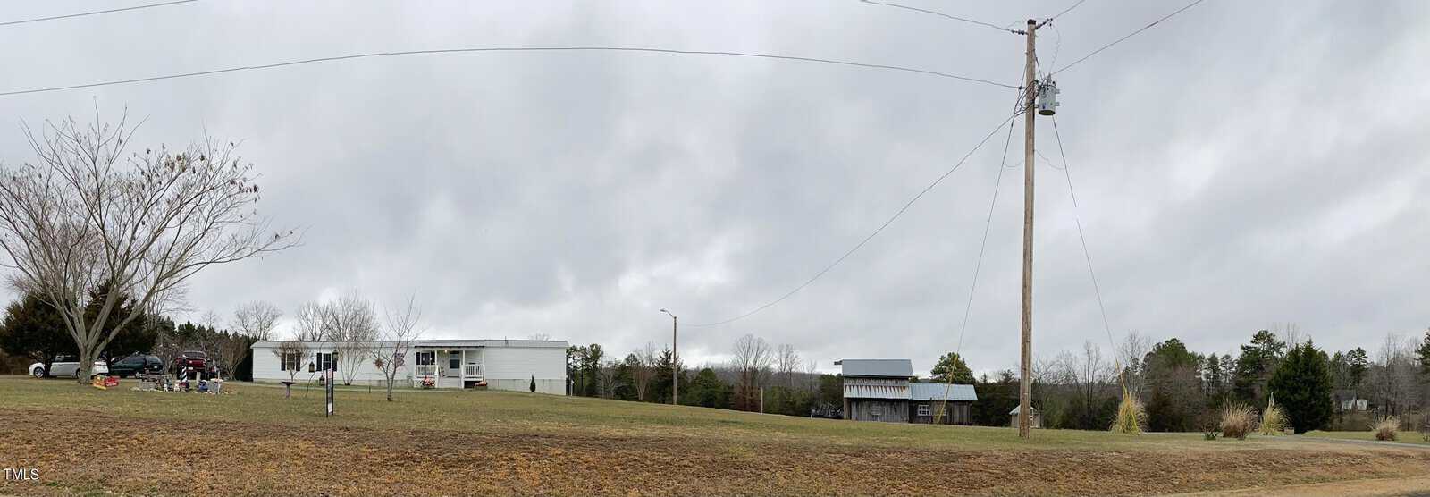 A rural scene shows a mobile home with cars parked nearby, surrounded by an expansive lawn. There's a barn in the background and a tall utility pole with wires stretching across the cloudy sky. Sparse trees dot the landscape.
