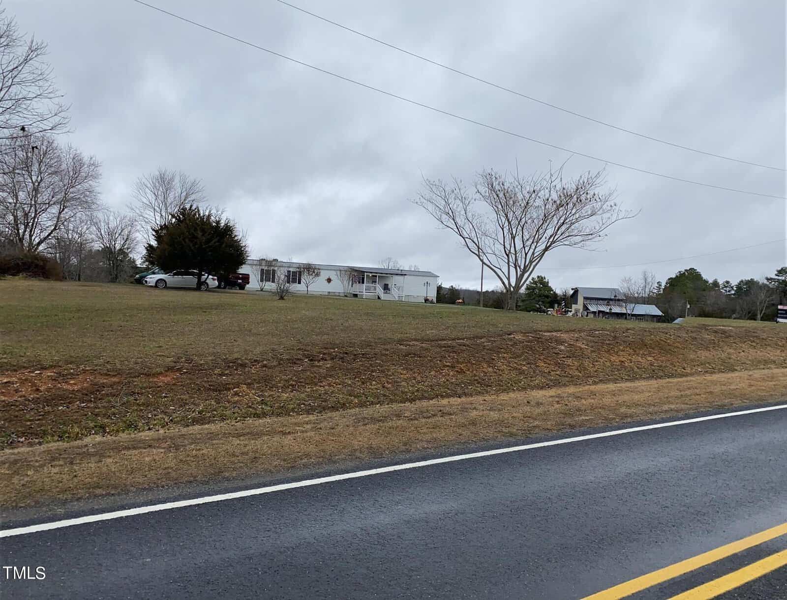 A rural scene with a cloudy sky, showing a long, single-story white building and a couple of cars on a grassy hill. Bare trees and a two-lane road are in the foreground.