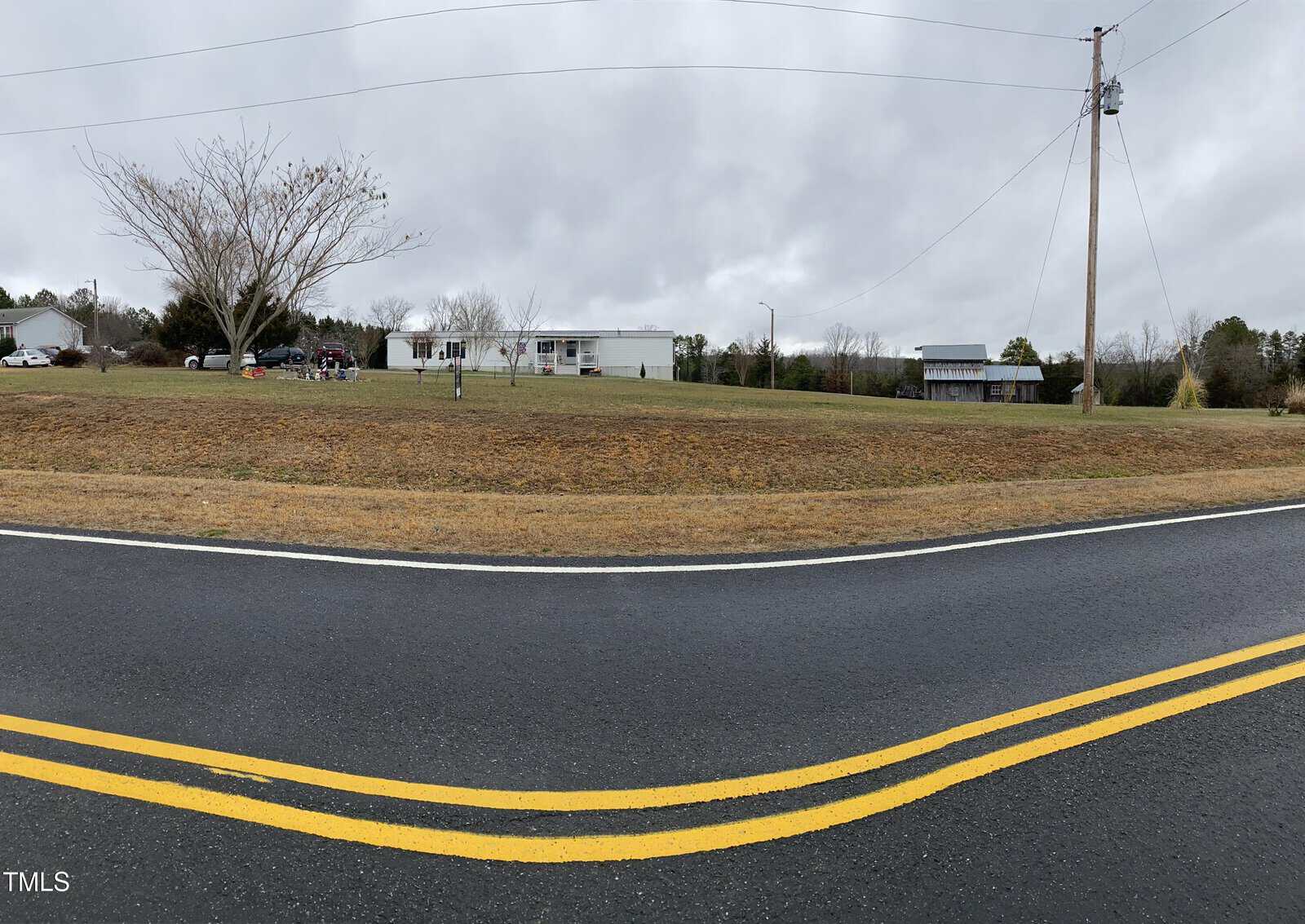 A panoramic view of a rural scene with a road in the foreground, a grassy lawn, and a house on a small hill. There are several trees, utility poles, and a shed under an overcast sky.
