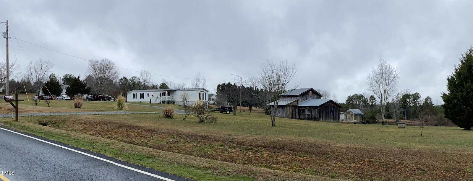 A rural landscape unfolds along 2637 Propus Wilkinson Road, featuring a road beside a grassy field. Several buildings, including a white mobile home and a wooden barn, dot the scene. Overcast skies and bare trees enhance the winter or early spring setting of this Bullock countryside.