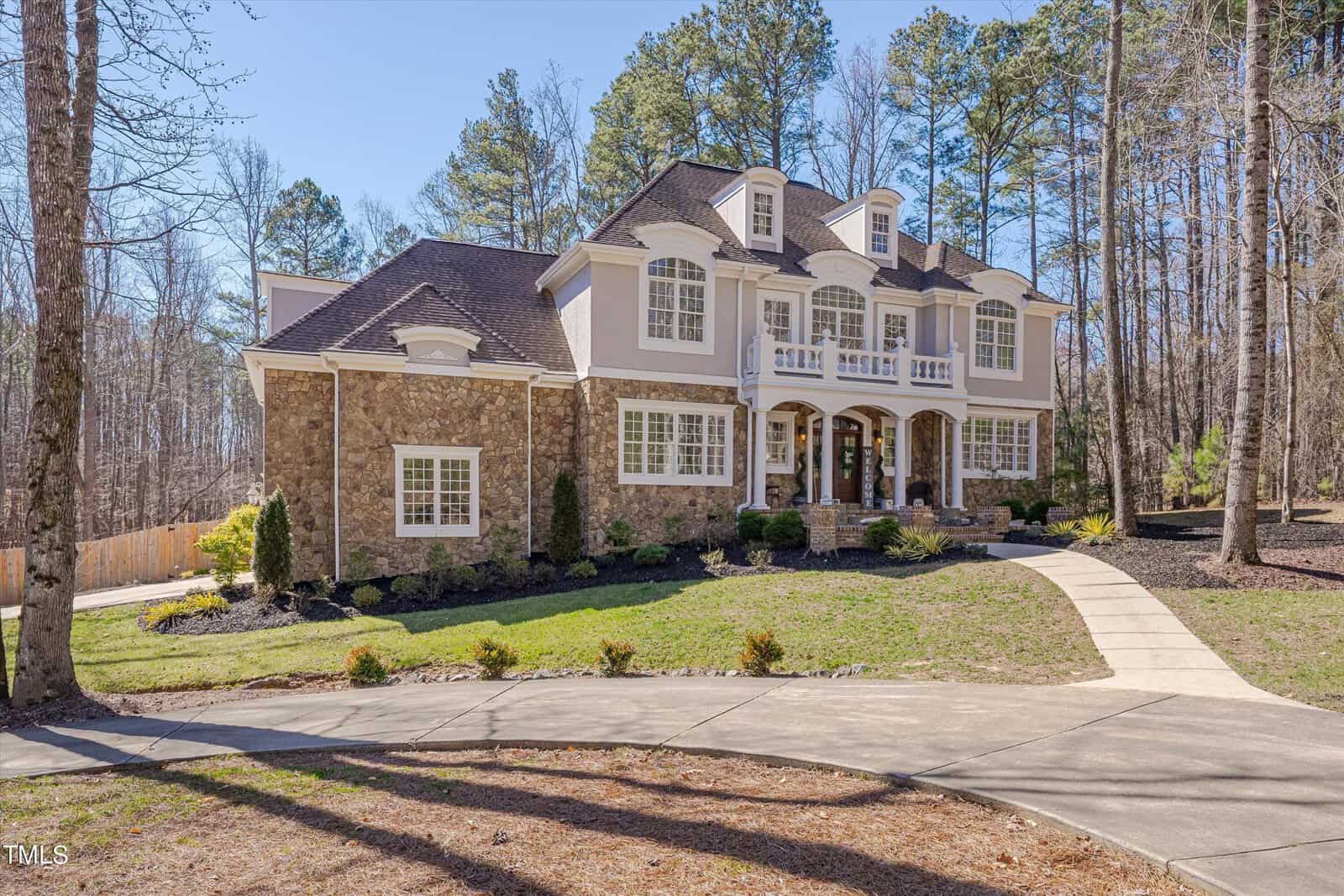 A large, elegant two-story house with a stone and stucco exterior stands surrounded by tall trees. It features a symmetrical design with multiple windows, a double front door, and a well-maintained lawn, connected by a curved pathway.