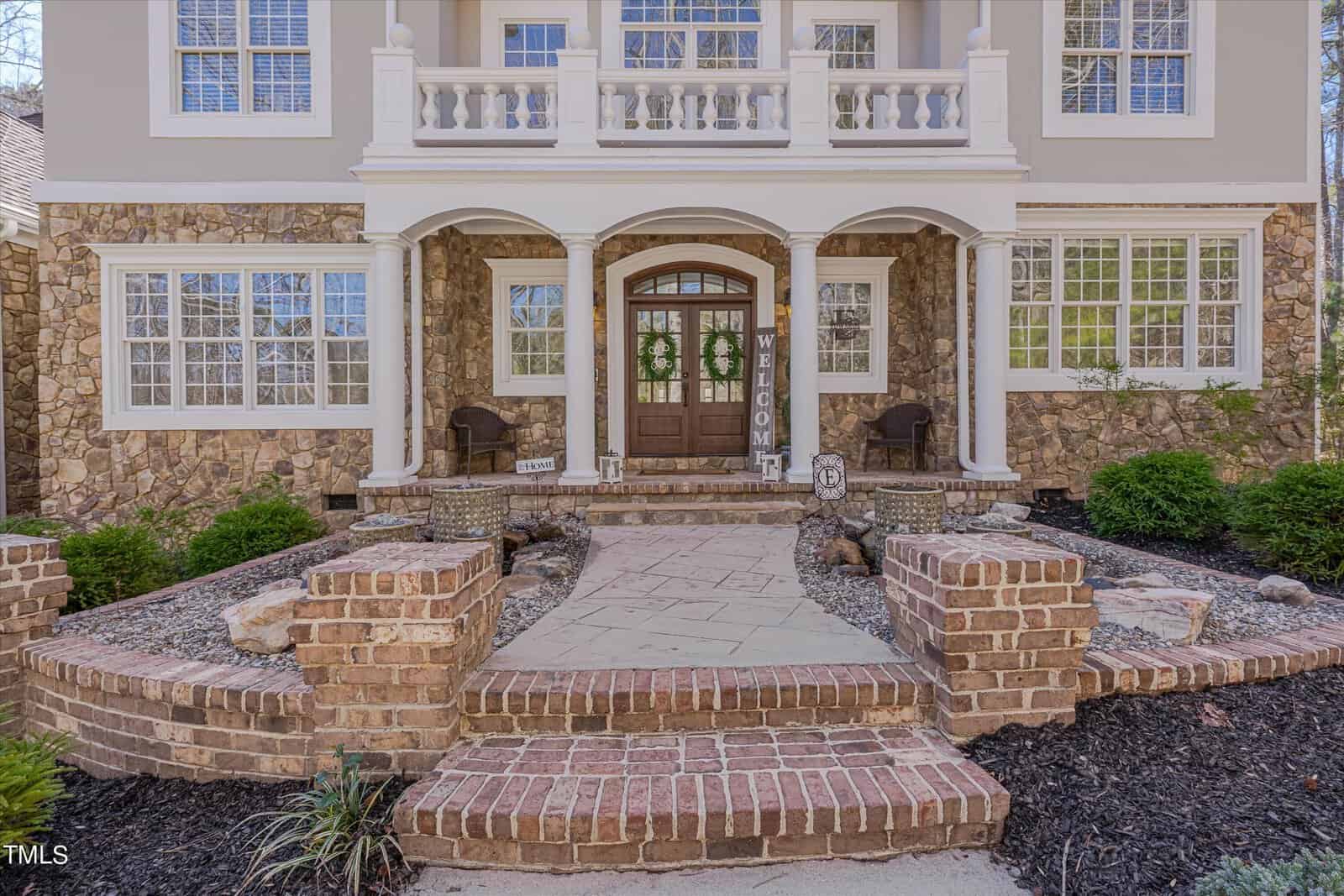 A grand entrance of a stone and stucco house featuring a double wooden door with wreaths. Brick steps lead to a porch adorned with columns and potted plants. Rock and mulch landscaping frames the pathway.