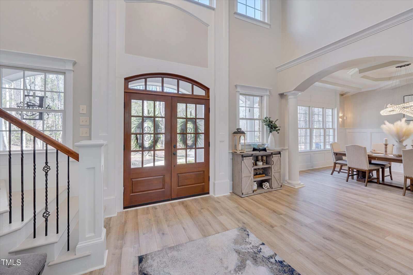 Spacious and bright entryway with a wooden double door and large windows. Stairs with iron railings are on the left, leading up. A dining area featuring a table and chairs is visible on the right. Light wood flooring throughout.
