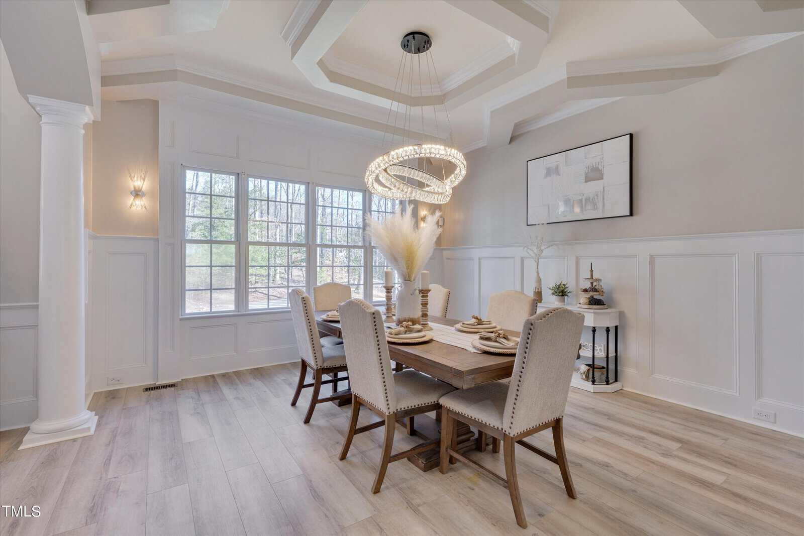 Elegant dining room with a wooden table set for six, featuring beige upholstered chairs and a modern chandelier. Large windows let in natural light, highlighting the light wood flooring and white paneled walls. A decorative piece hangs on the wall.