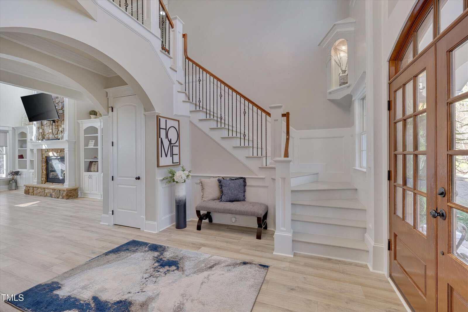 Spacious entryway with a wooden staircase, blue and white rug, bench with pillows, and a "Home" sign. Large windows and a stone fireplace are visible in the adjacent living area. Light wooden flooring enhances the airy atmosphere.