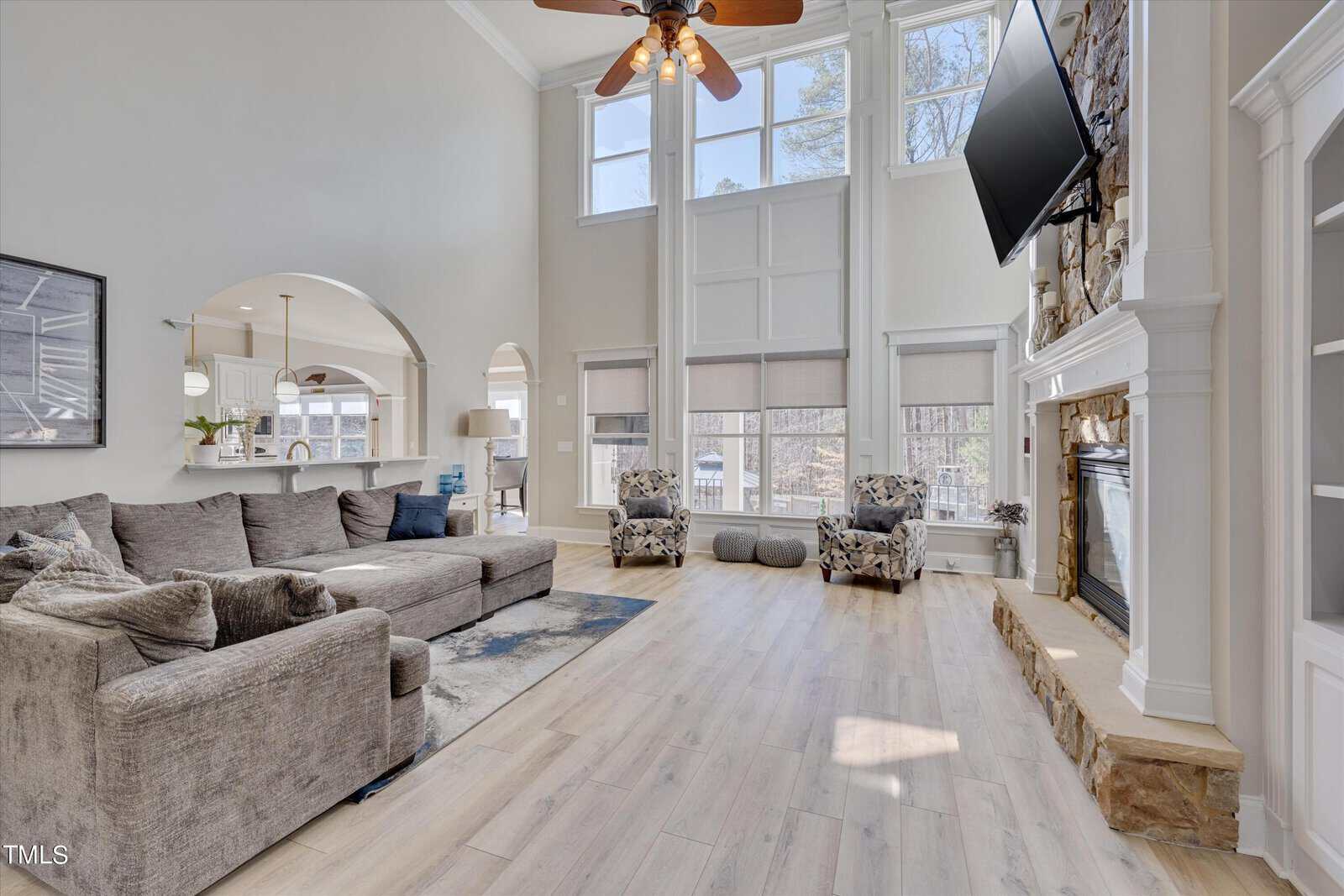 Spacious living room with high ceilings, a large sectional sofa, and two patterned armchairs. Features a stone fireplace with a mounted TV and large windows allowing natural light. Neutral tones and wooden flooring add warmth to the modern decor.