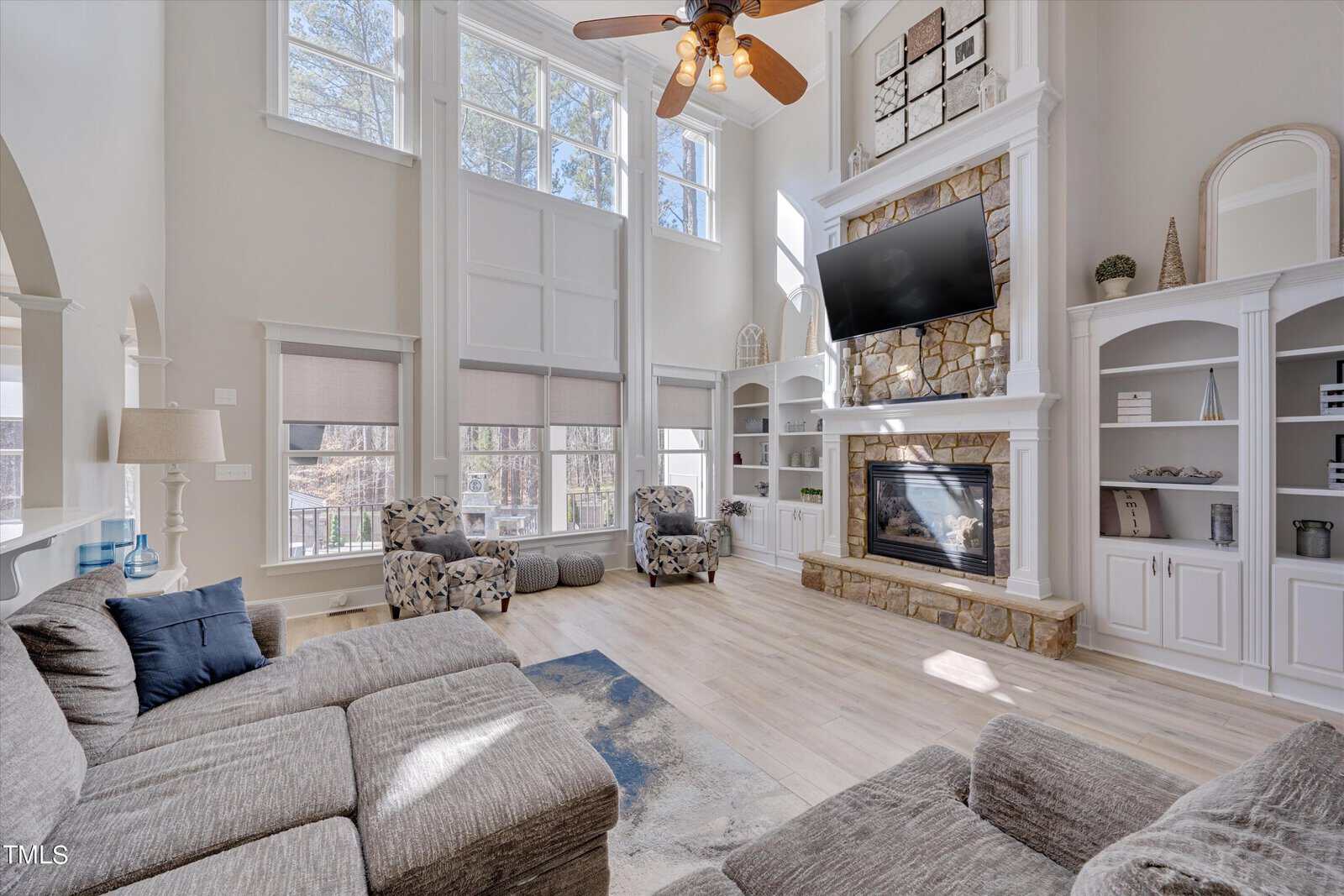 A spacious, sunlit living room with tall windows, a stone fireplace, and a mounted TV. It features a gray sectional sofa, patterned armchairs, and built-in white shelves. A ceiling fan hangs above, and the room has light wood flooring.