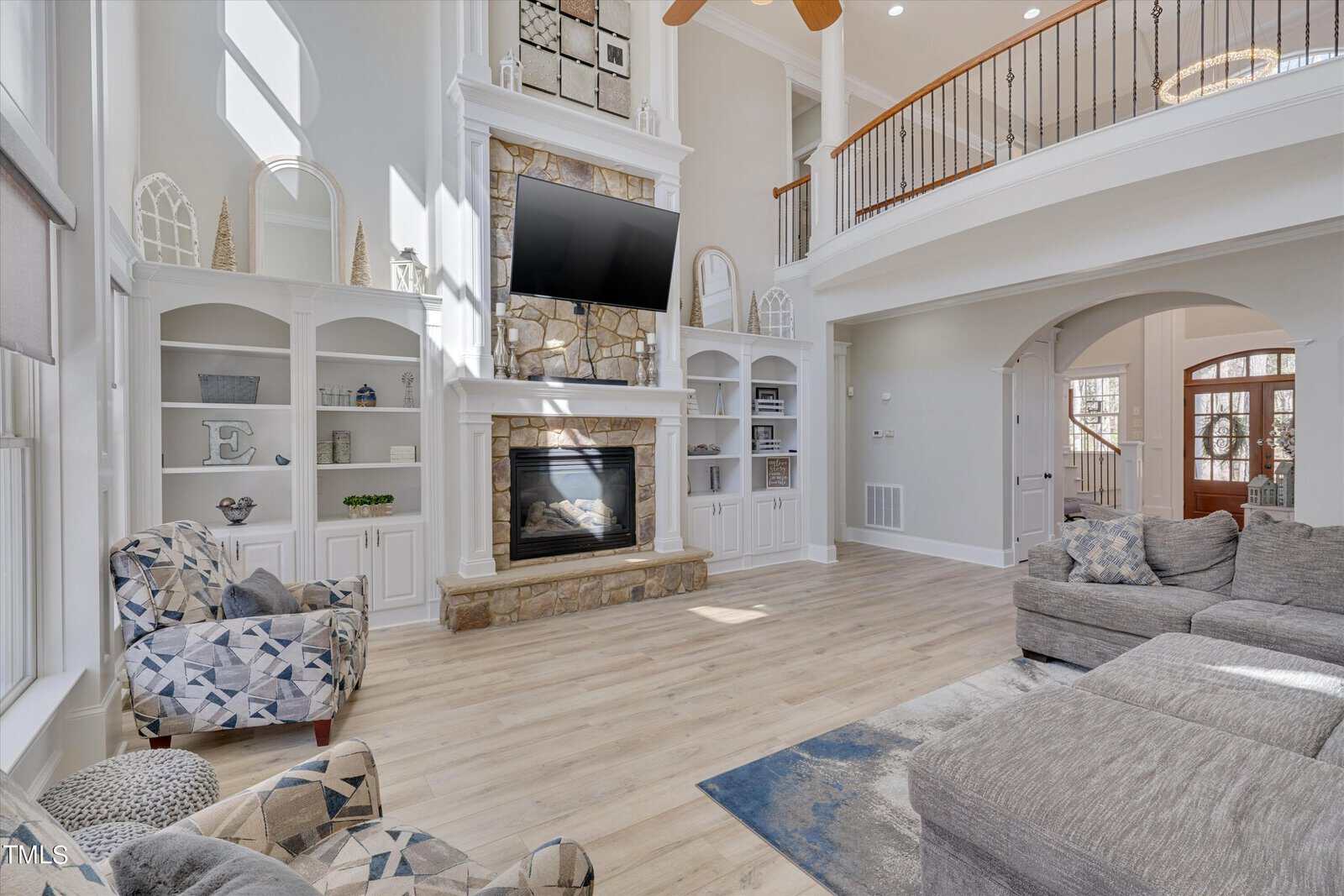 A spacious living room with a tall stone fireplace, a mounted TV, and built-in shelves filled with decor. A large sectional sofa and patterned armchairs create a cozy seating area. Bright natural light streams in from large windows above.