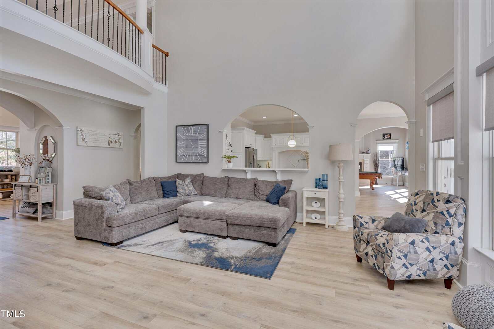 Spacious living room with gray sectional sofa and blue patterned armchair. Light wood flooring and a large area rug enhance the space. Arched openings lead to a kitchen with white cabinetry. Ample natural light fills the room through large windows.