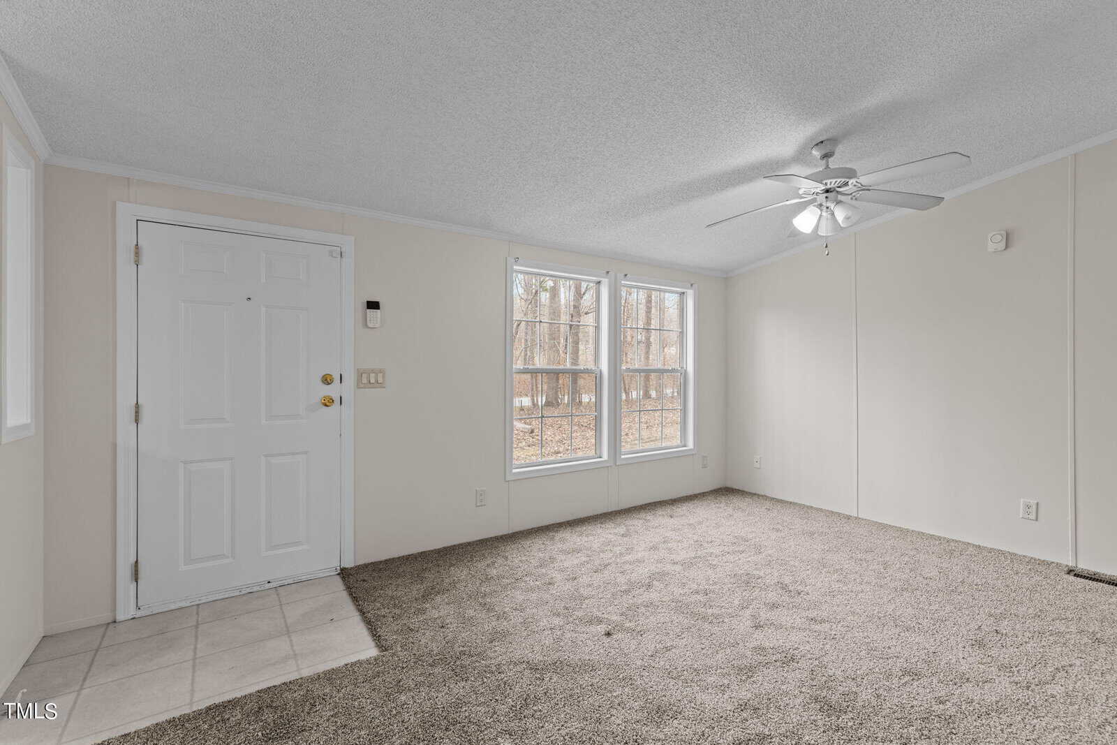 A minimalist room featuring light beige walls and carpet, with a white front door on the left and two large windows with views of a wooded area. A white ceiling fan with light is centrally mounted on the ceiling.