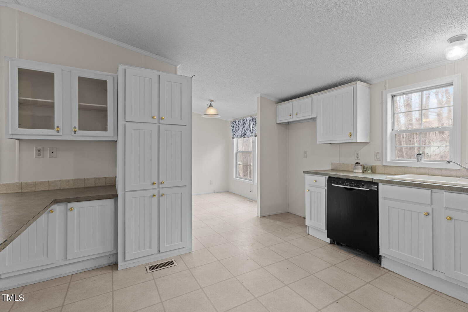 A spacious kitchen features white cabinets, a black dishwasher, and beige countertops. A sunlit dining area is visible in the background. The kitchen has tile flooring and a light fixture hanging from the ceiling in the dining area.