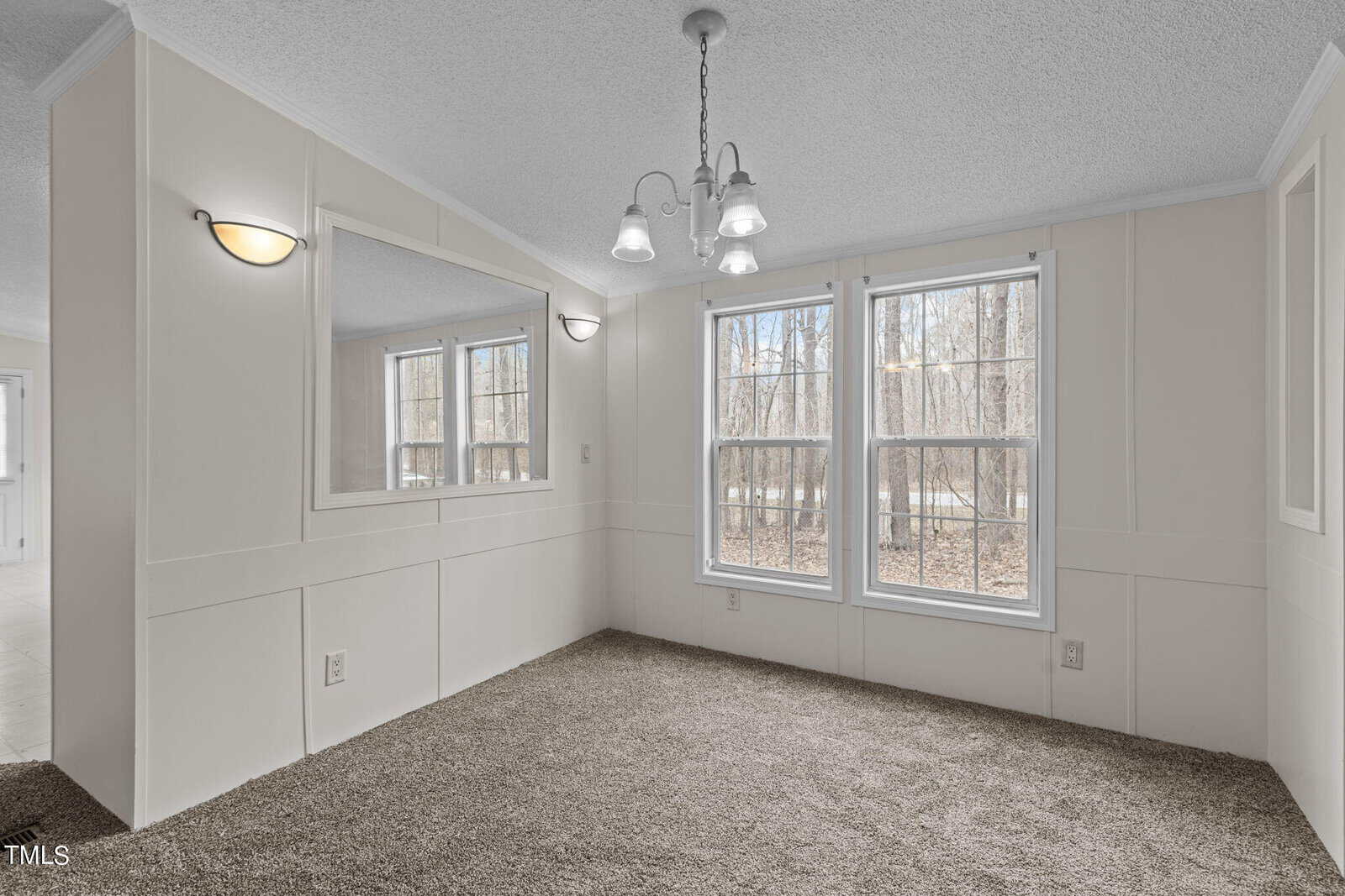 A bright room with beige carpet, white wainscoting walls, two large windows overlooking a wooded area, a wall-mounted mirror, and a ceiling light fixture with three lamps. The room connects to other areas through doorways visible on the left.