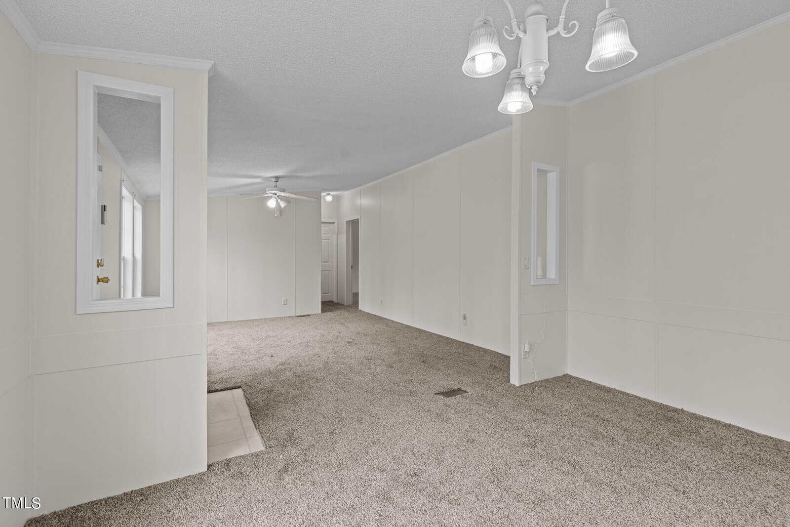 A spacious, empty living room with beige carpet and white walls. The room features two small, vertical windows with white trim on an interior wall adding to the brightness of the space. A ceiling fan and a white chandelier are mounted on the ceiling.