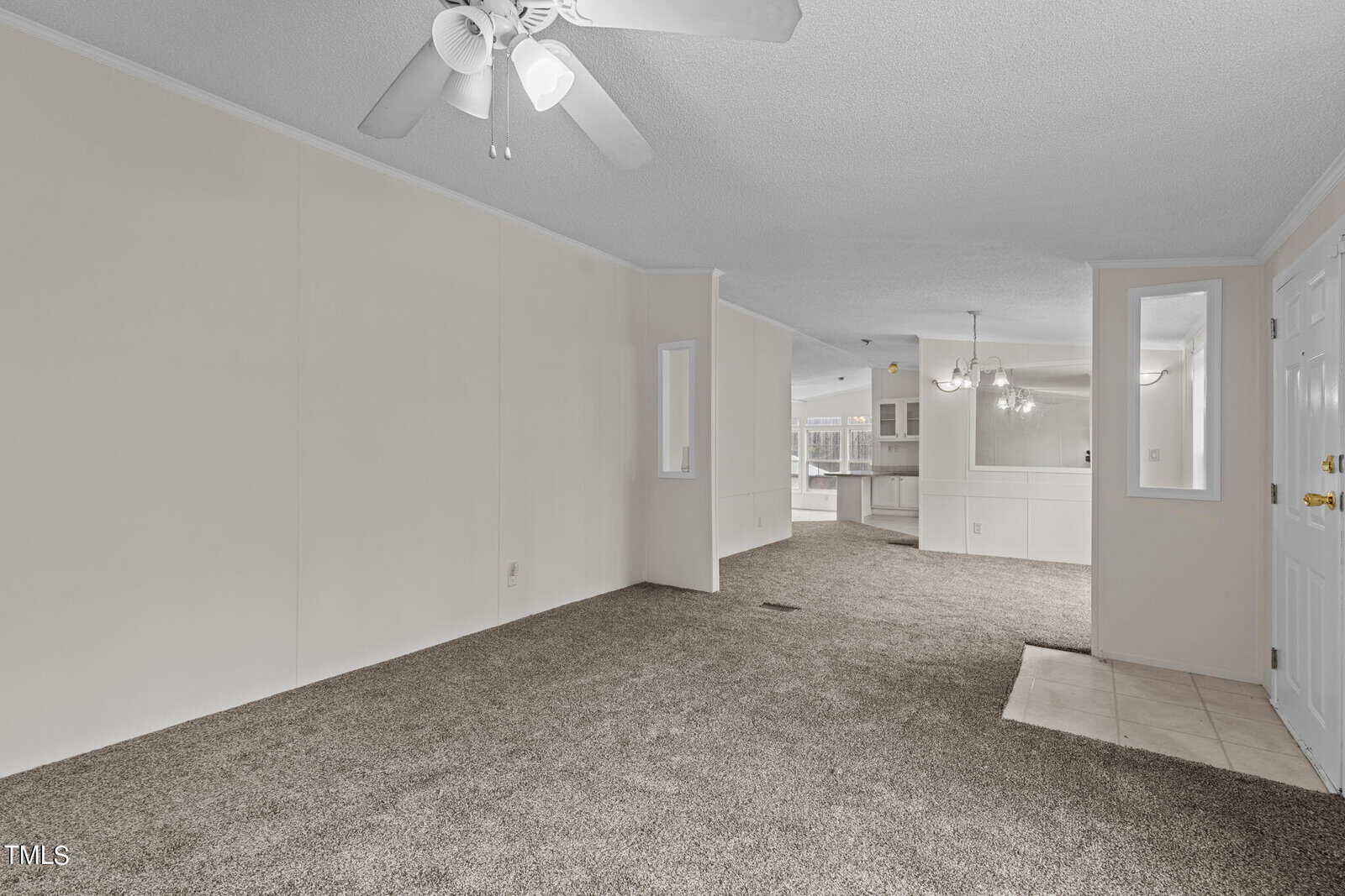 A spacious living area with light gray walls, gray carpet, and a white ceiling fan. Adjacent to it is an open dining area with a chandelier and kitchen with white cabinets partially visible in the background. Natural light enters from windows in the distance.