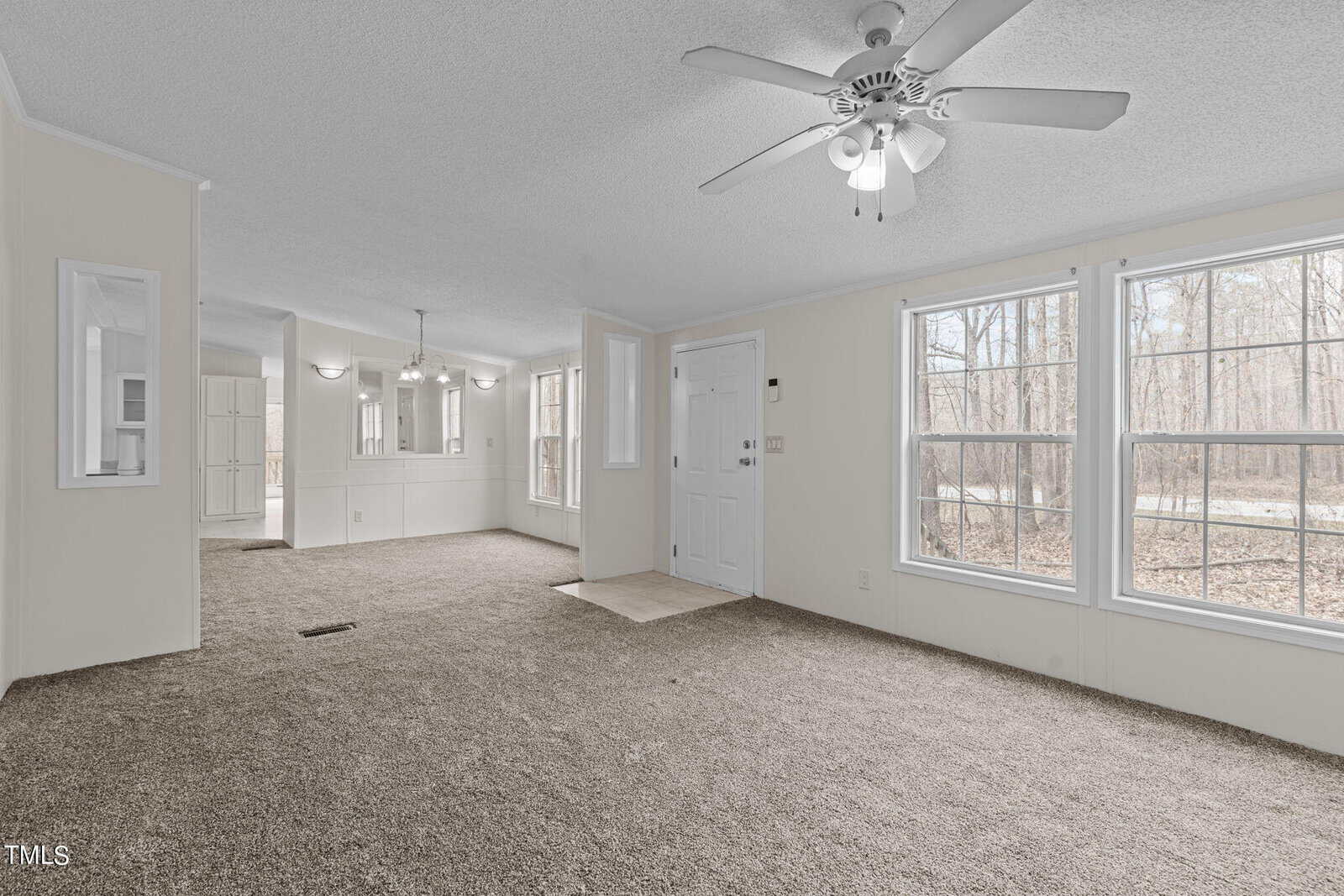 A spacious, empty living room with beige carpet and a textured white ceiling featuring a ceiling fan with lights. Large windows on the right bring in natural light, and an open doorway leads to a dining area. A white door is visible near the center back wall.