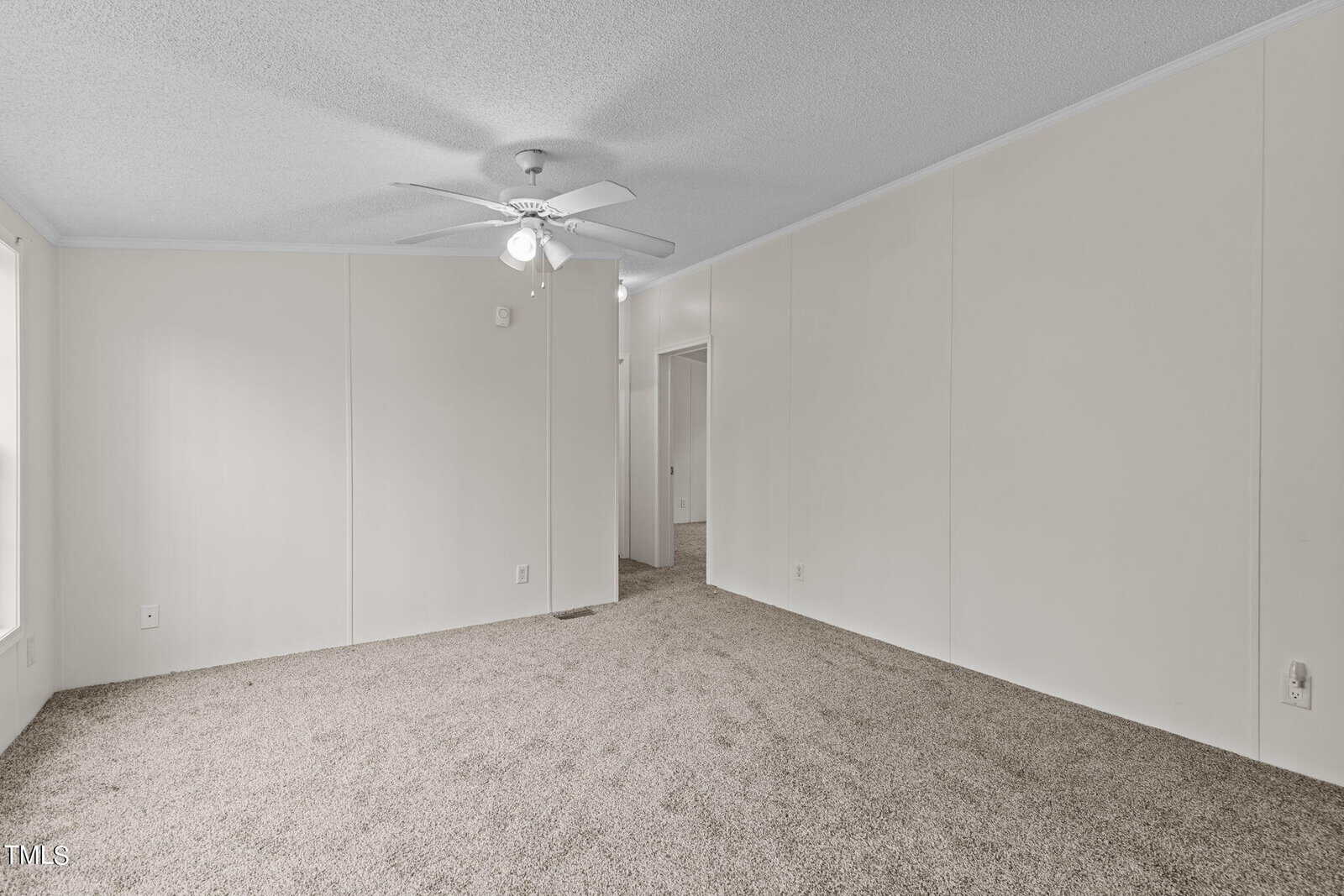A spacious and empty room with beige carpet flooring and white walls. A ceiling fan with lights is mounted at the center of the ceiling. There is a doorway leading to another room and a large window allowing natural light to enter.