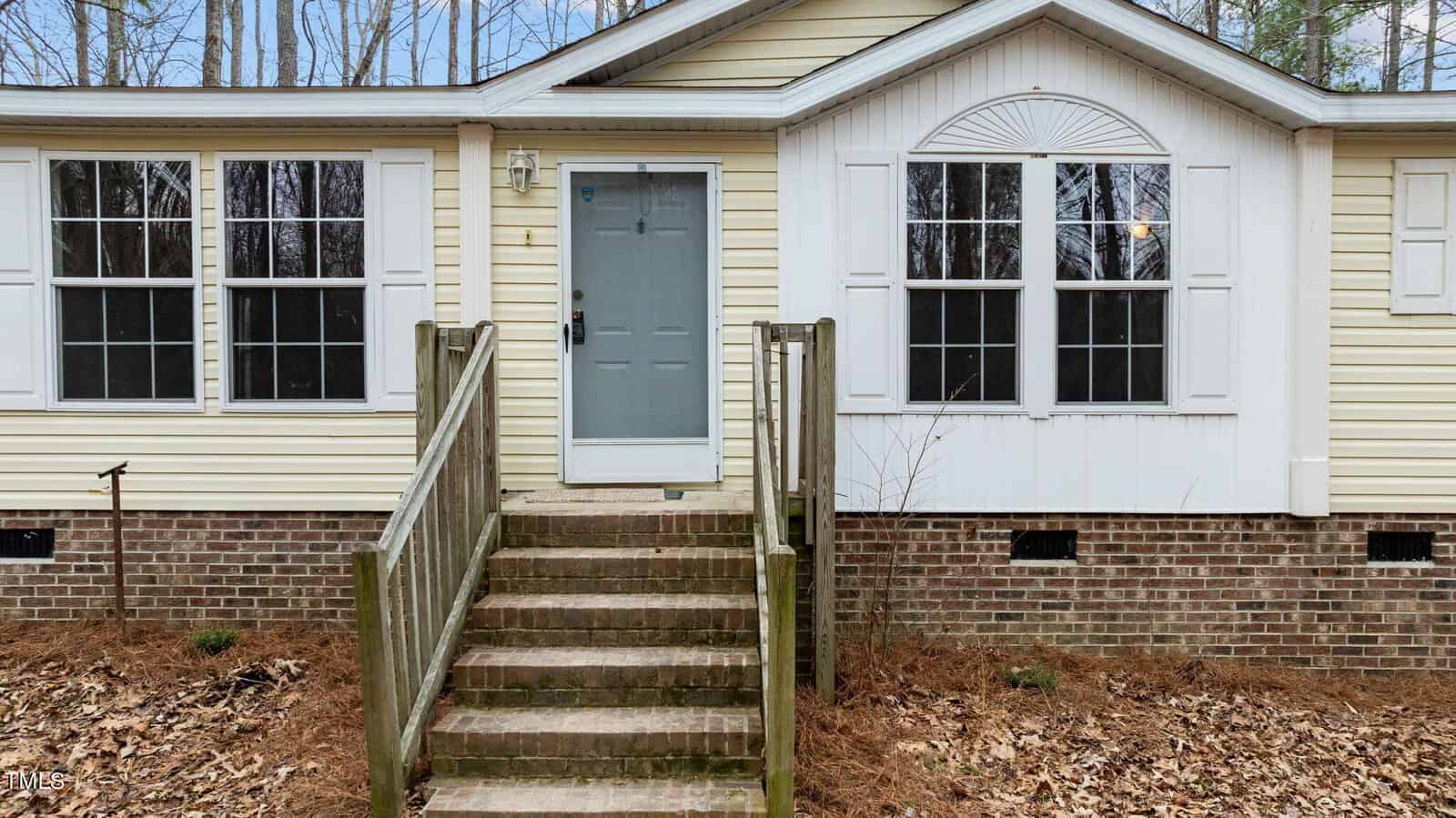 A beige, single-story house with white trim and a gray front door, located at 1188 Sims Bridge Road, Kittrell. The entrance features a short set of brick steps and a railing. Tall windows flank the door, and the house is surrounded by a wooded area with fallen leaves on the ground.