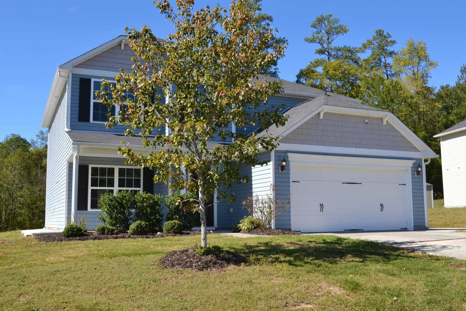A two-story suburban house with light blue siding and a white garage door. There's a small tree in the front yard and a manicured lawn. The sky is clear and sunny.