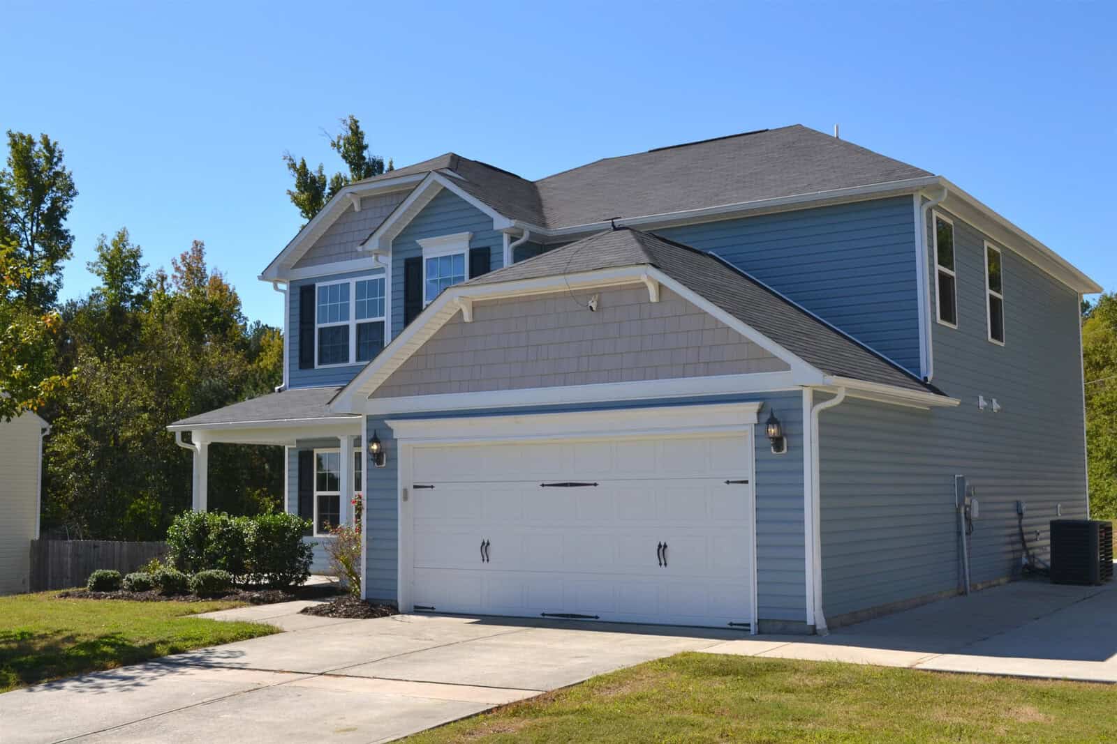 A two-story suburban house with blue siding, a white garage door, and a small front porch. The driveway and lawn are neatly maintained, and trees are visible in the background under a clear blue sky.
