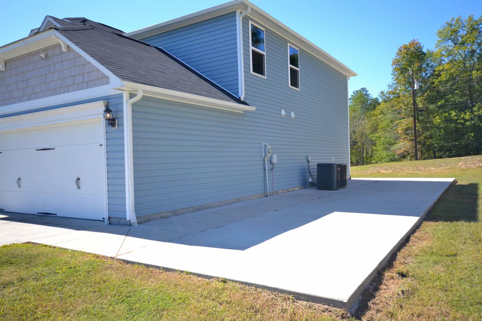 Side view of a two-story blue house with white trim and a black roof. The house features a double garage door and a large concrete driveway. Trees and grass surround the property under a clear blue sky.