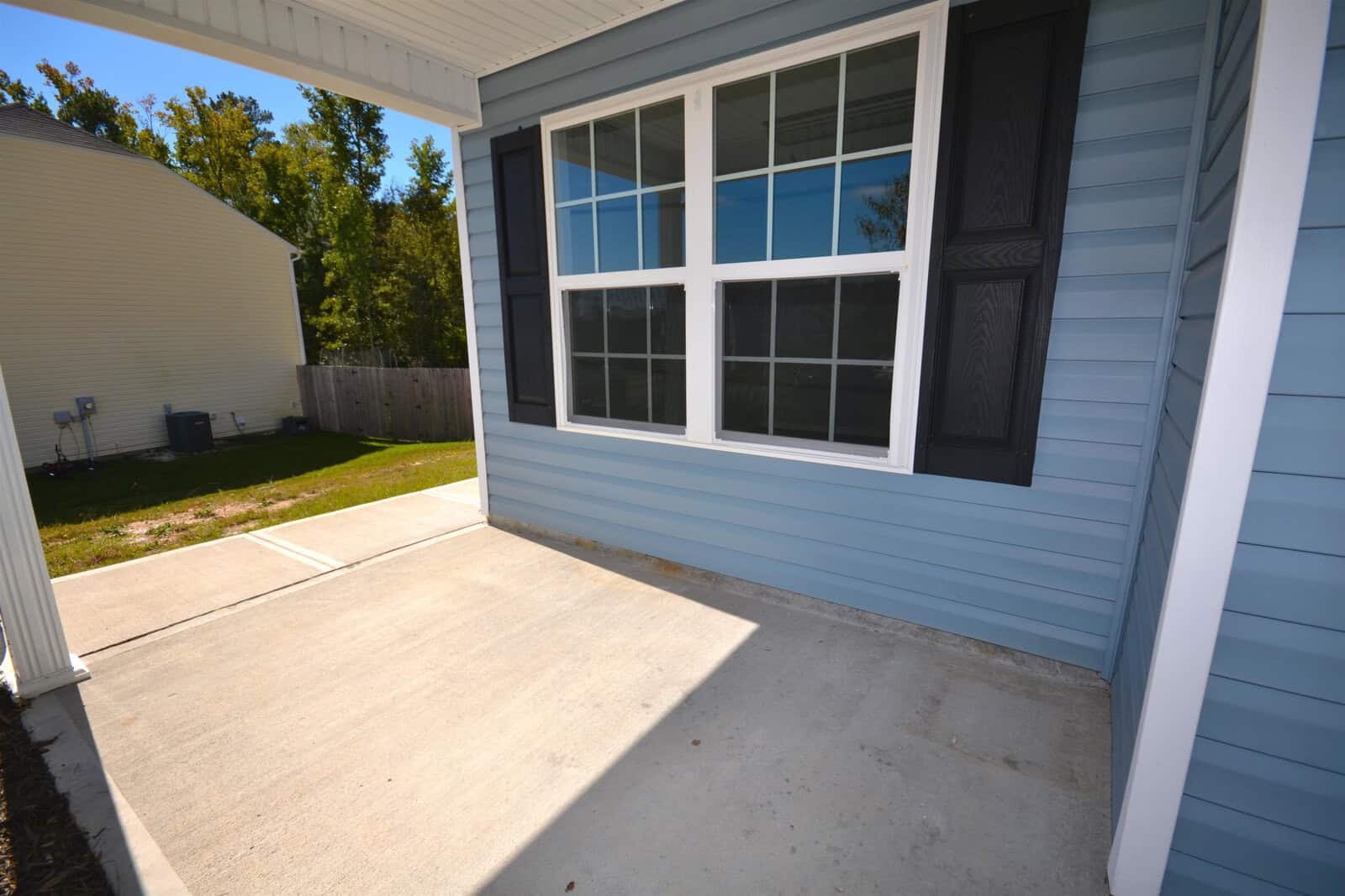 A house exterior with a concrete patio. The siding is blue with white trim and features a large window with black shutters. A concrete path leads toward a lawn and fence, and a neighboring house is visible in the background.