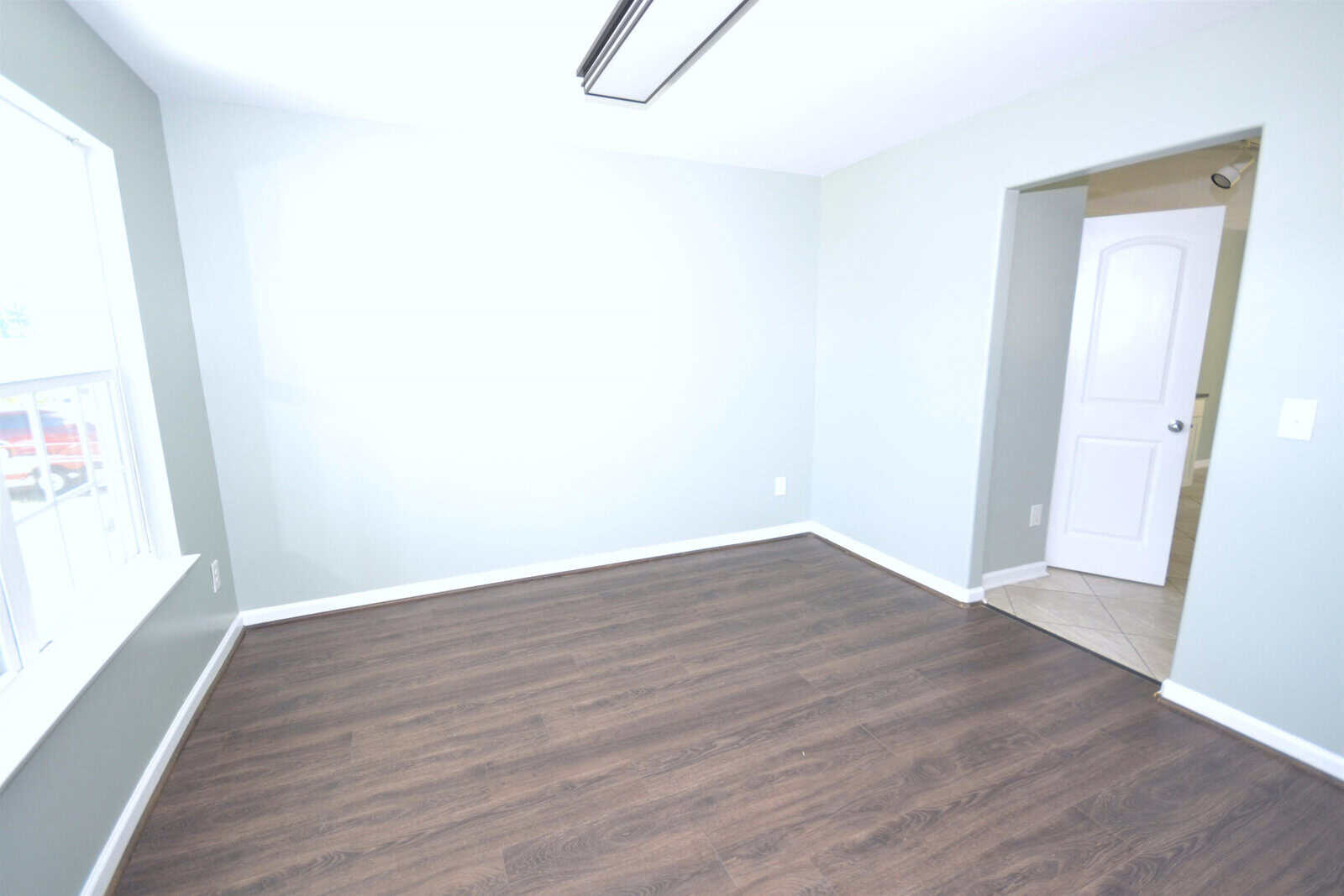 An empty room with light gray walls and dark wood flooring. A large window is on the left, and an open doorway with a white door leads to another room. A fluorescent light fixture is on the ceiling.