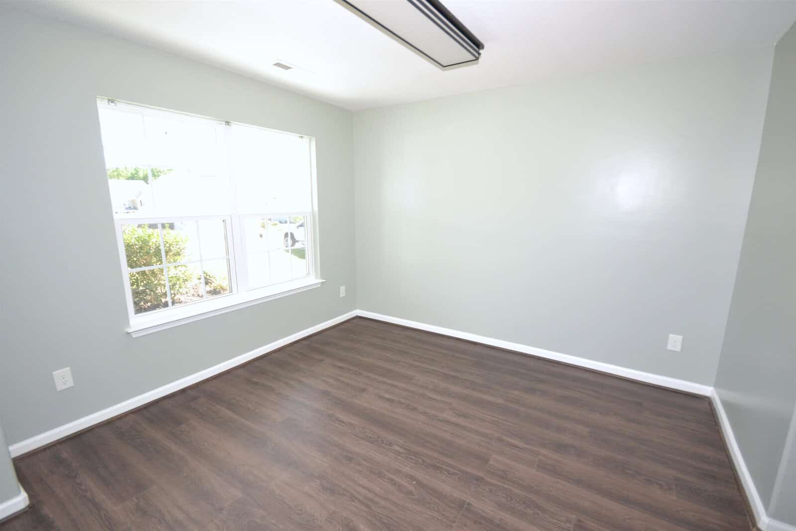 An empty room with pale green walls, a large window with a view of greenery, and dark wooden flooring. The ceiling has a fluorescent light fixture. The room is bright with natural light.