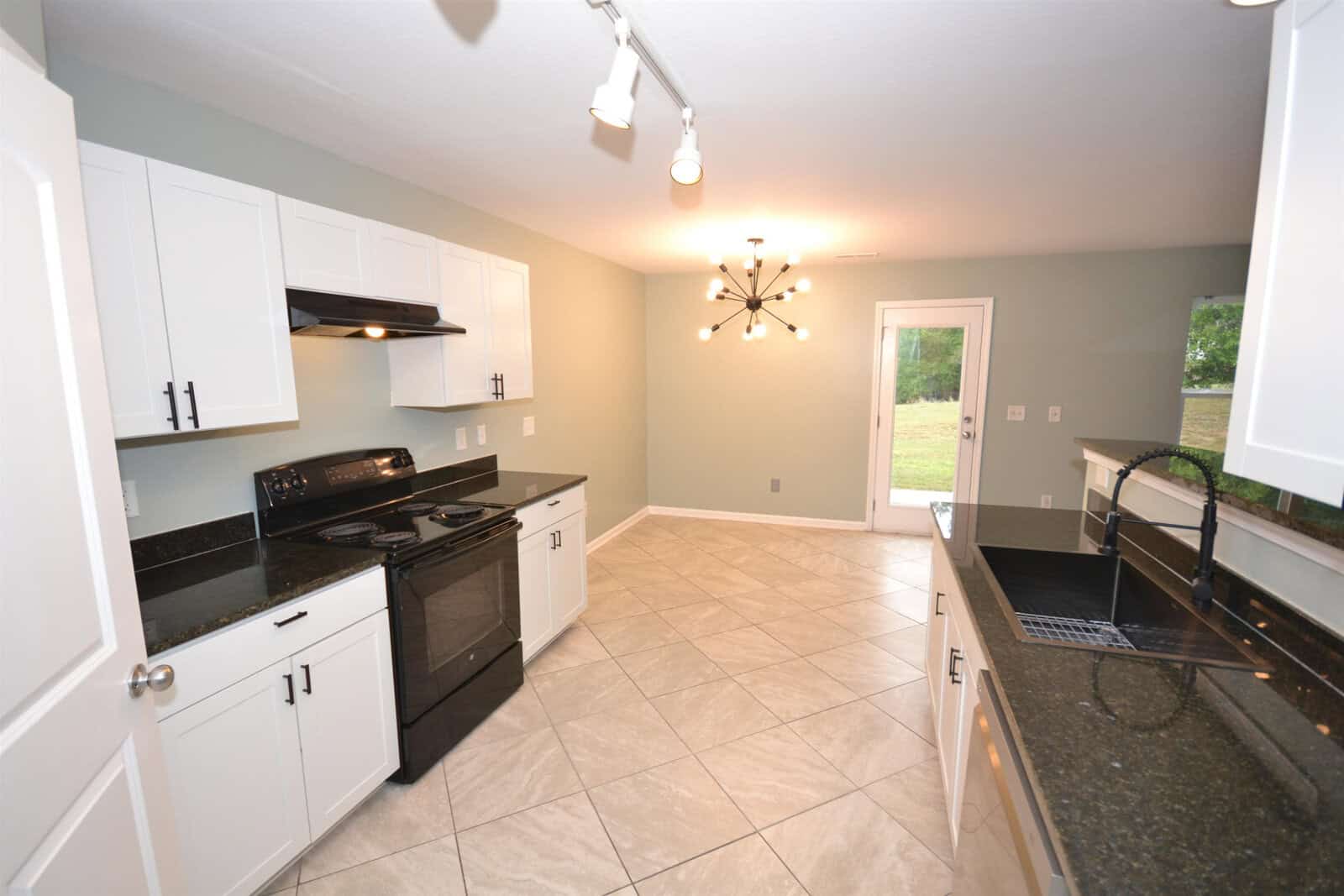 A modern kitchen with white cabinets, black countertops, and tile flooring. It features a black stove and hood, a sink, and a contemporary ceiling light fixture. There is a window and a glass door leading outside, allowing natural light to enter.
