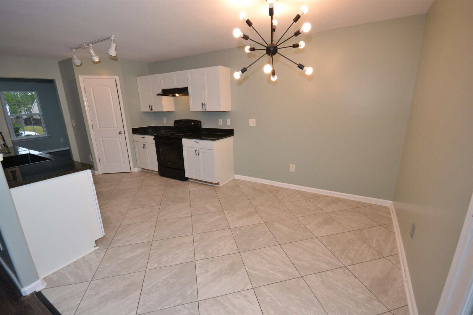 A modern kitchen with light green walls, black granite countertops, white cabinets, and a black stove. A unique chandelier with exposed bulbs illuminates the room. The floor is tiled in a beige pattern. A door leads to an adjacent room.