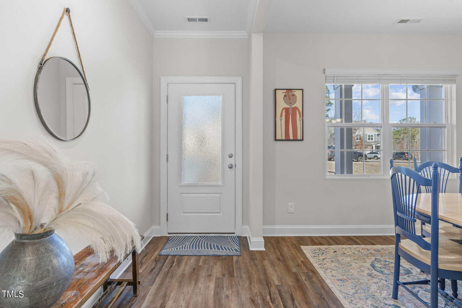 Bright entryway with a white door, round mirror above a wooden bench with a vase of pampas grass, artwork on the wall, and a window letting in natural light near a dining table with blue chairs.