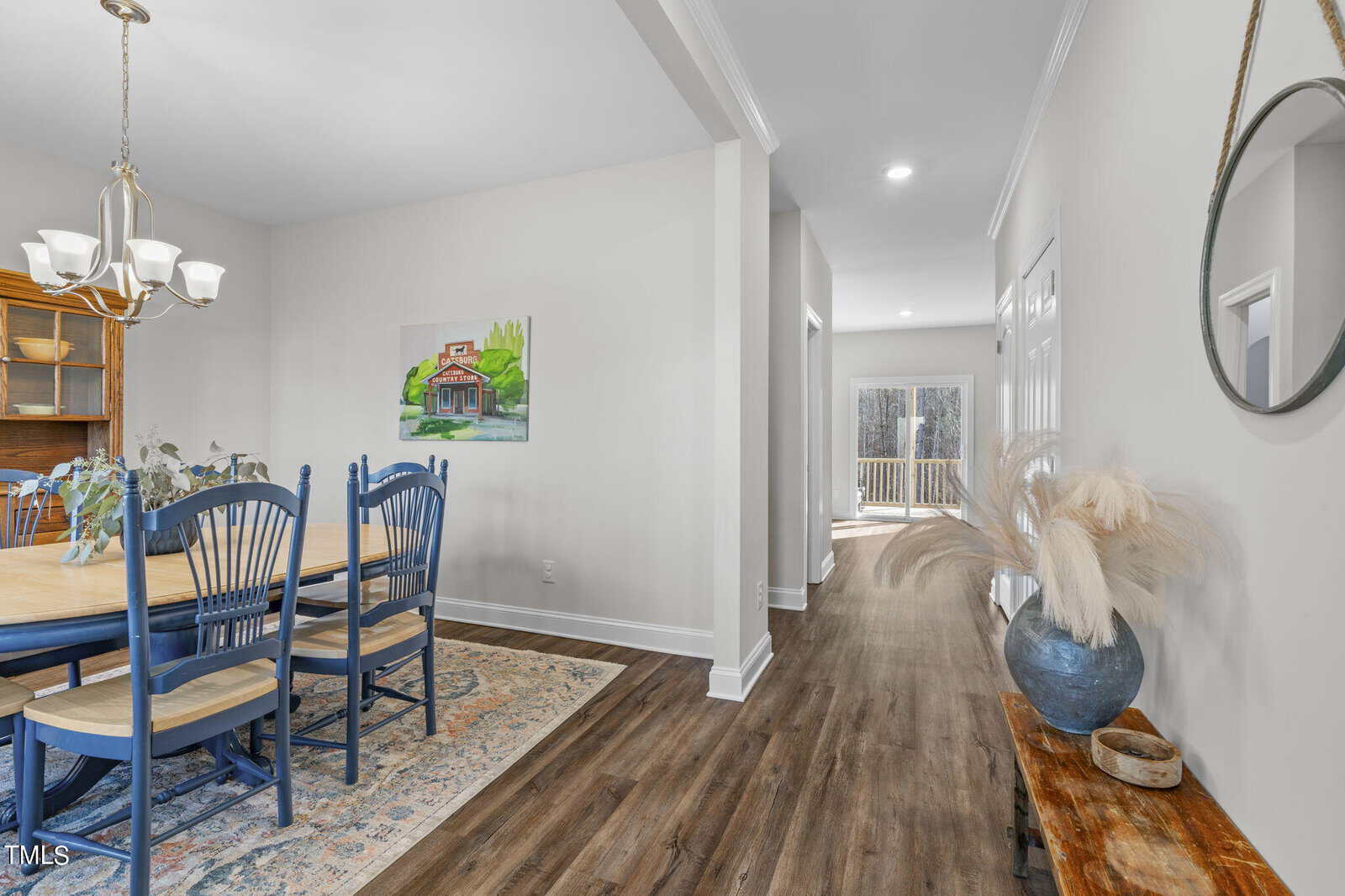 A bright dining area with a wooden table and blue chairs sits beside a hallway with wood flooring. A round mirror and vase with pampas grass are on a bench. Sunlight streams in from glass doors at the end of the hall.
