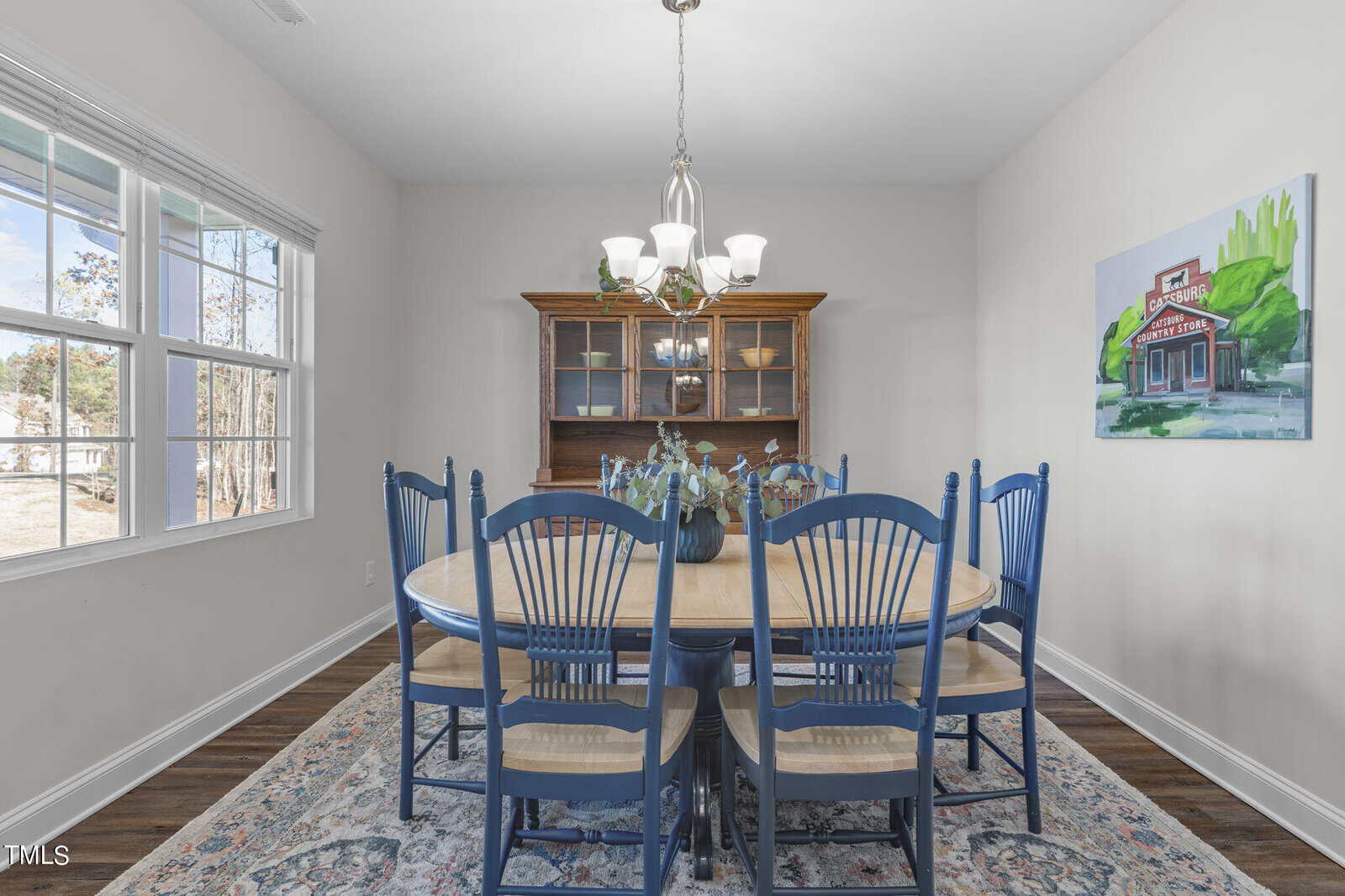 A bright dining room with a wooden table, eight blue chairs, a patterned rug, a chandelier, a wooden hutch, a plant centerpiece, wall art, and large windows letting in natural light.