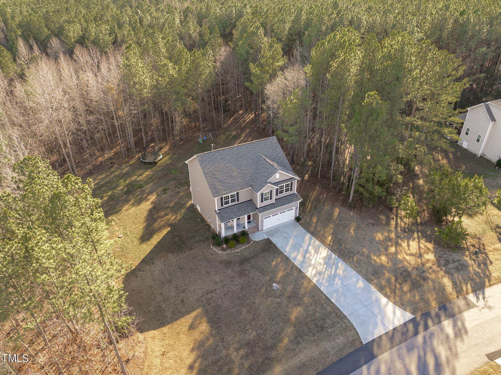 Aerial view of a two-story house with a gray roof and attached garage, surrounded by a large yard and dense forest, with a long driveway leading to the road.