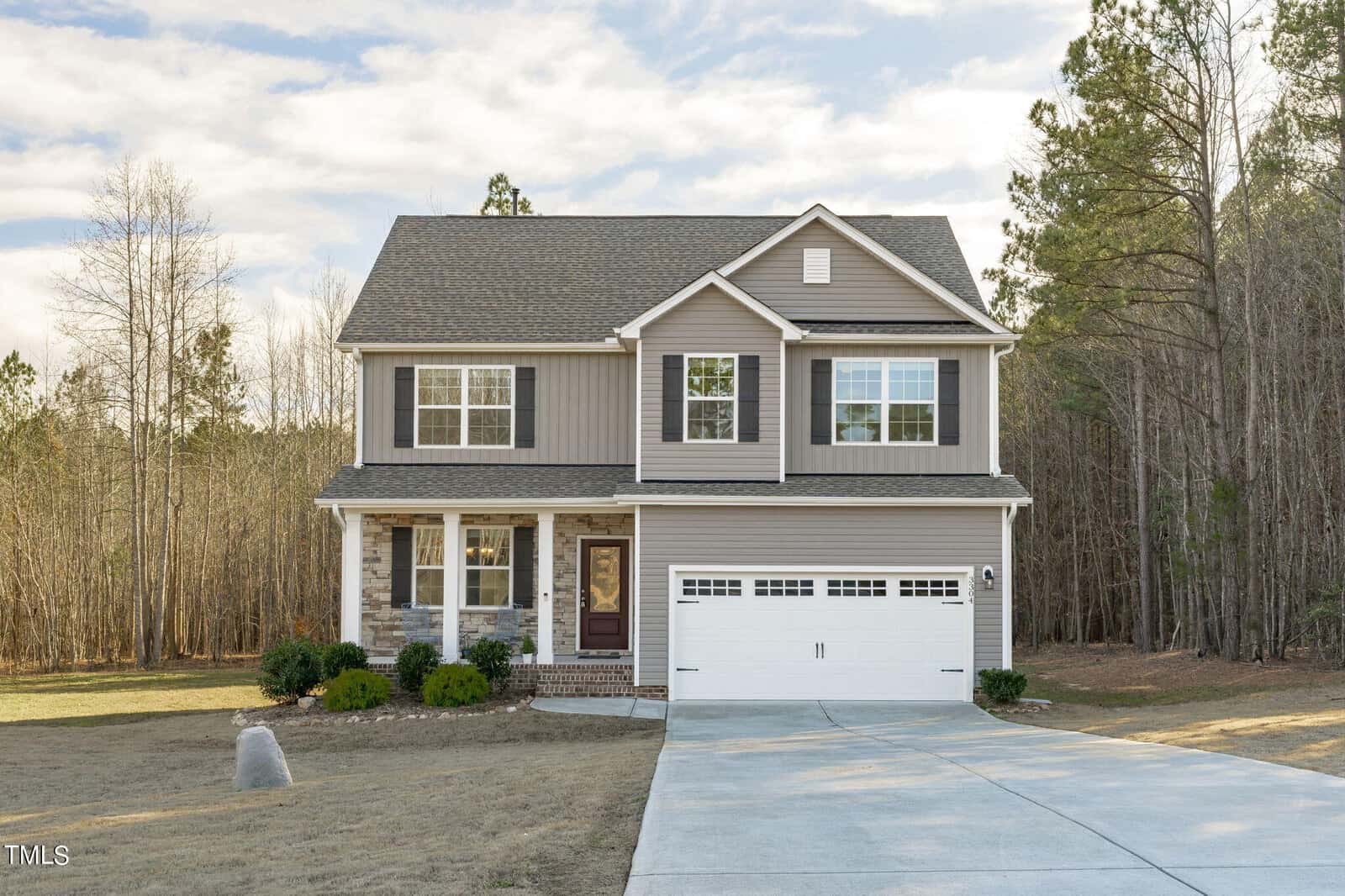 Charming two-story suburban home at 3304 Wiltshire Way in Creedmoor, featuring beige siding, stone accents, a covered front porch, double garage, and driveway. Surrounded by trees and bushes with an open sky overhead.