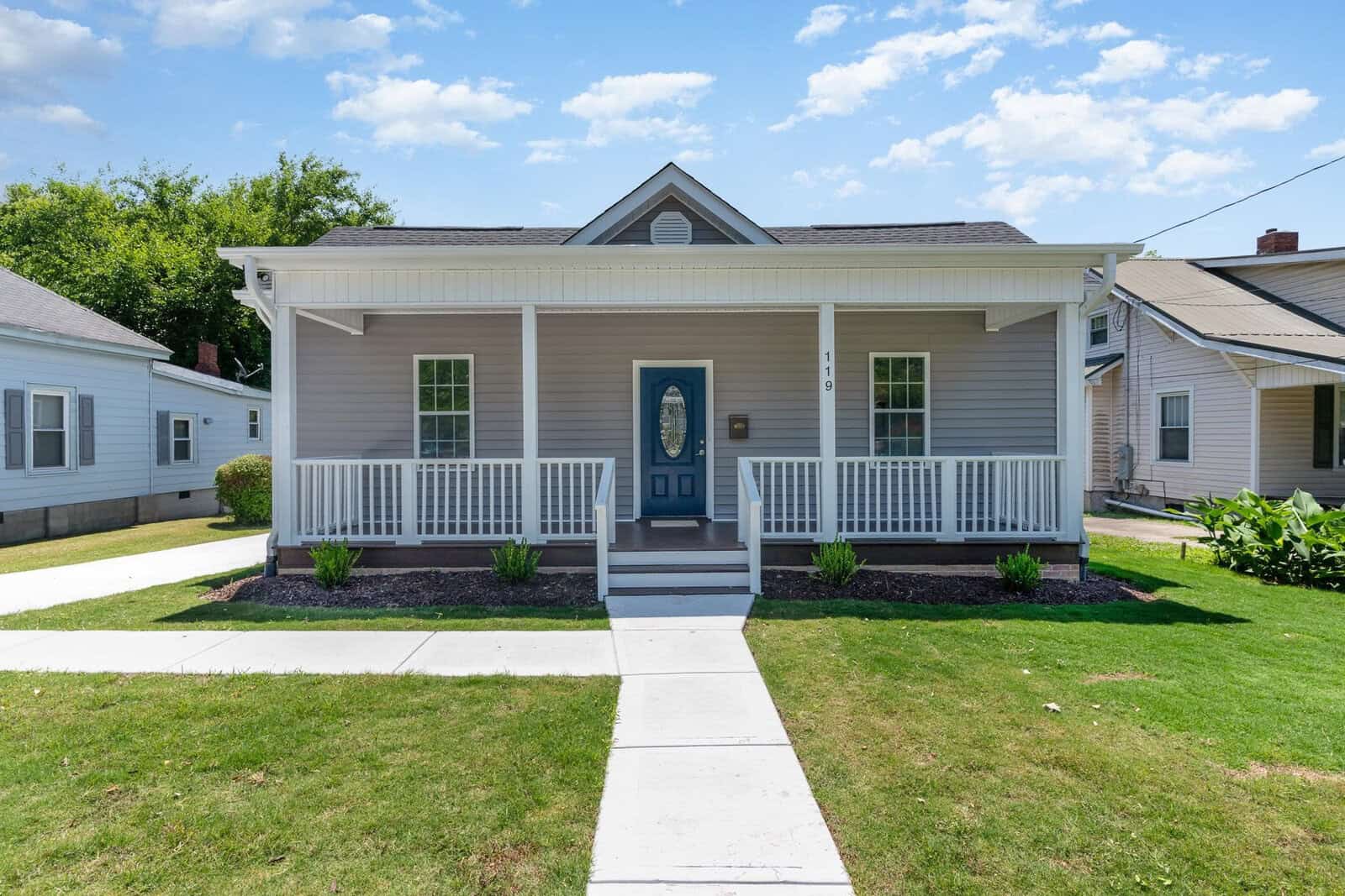 A small, single-story house at 119 Sycamore Street features a gray exterior with a charming blue front door. The Oxford-style covered porch boasts white railings, while a walkway meanders through the lush grassy lawn beneath a clear sky with a few clouds.