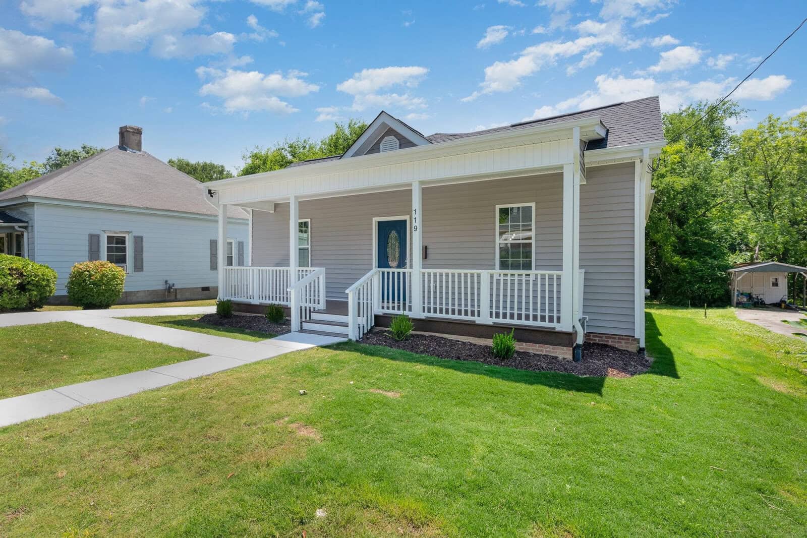 A gray house with a blue front door and white trim. It features a covered porch with white railings. The yard is grassy, and the sky is partly cloudy. Another house and a driveway are visible on the left.