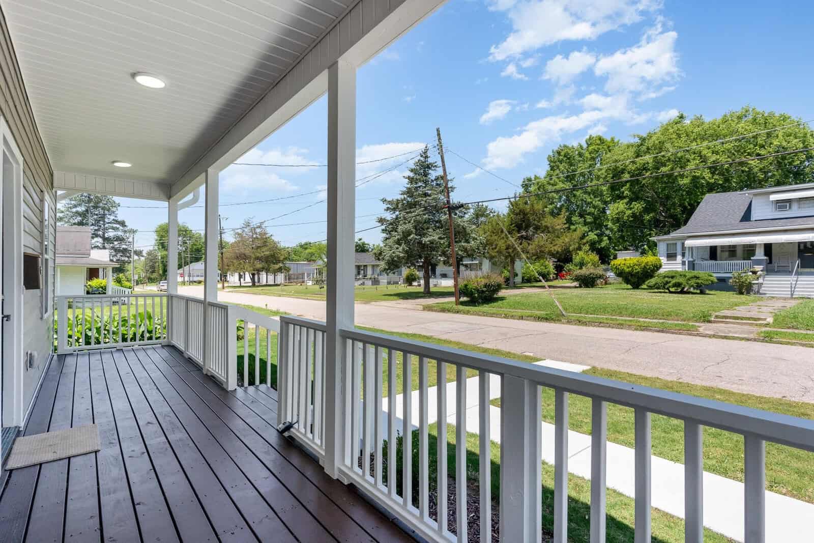 A view from a wooden porch with a white railing, overlooking a suburban neighborhood street. There are trees, a clear blue sky, and houses with lawns on both sides of the street.