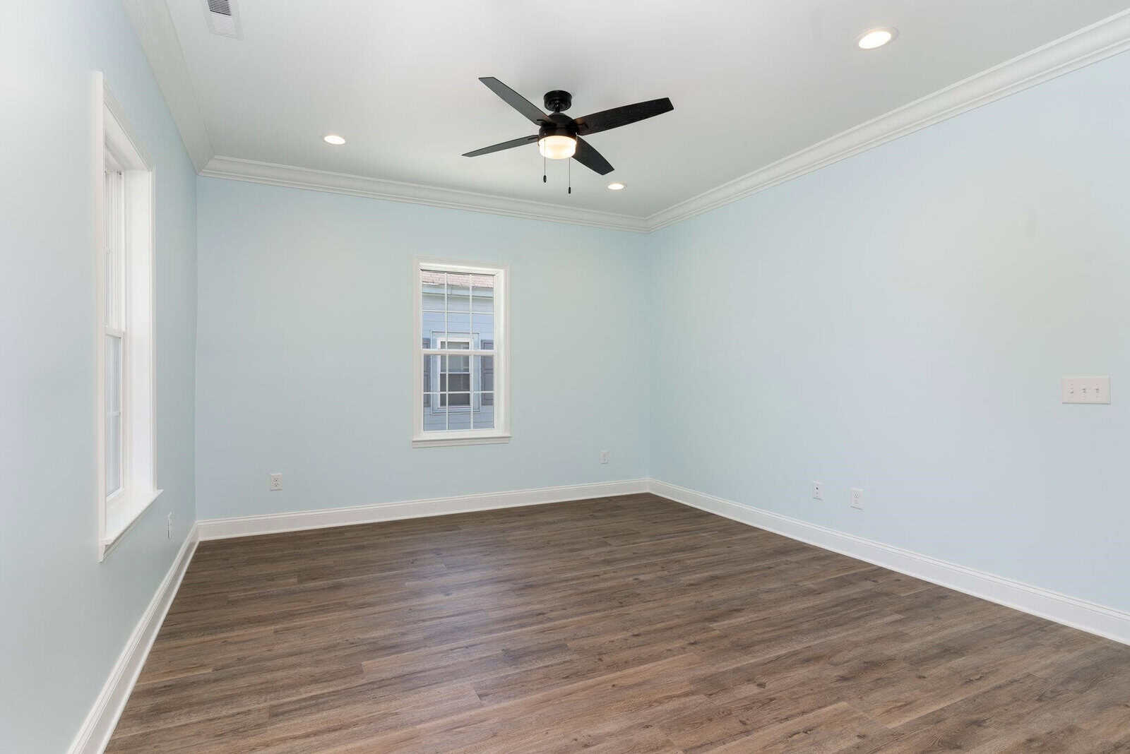 A spacious, empty room with light blue walls and dark wood flooring. It features a central window with white trim and a black ceiling fan with lights. Recessed lighting is installed in the ceiling, and there's ample natural light.