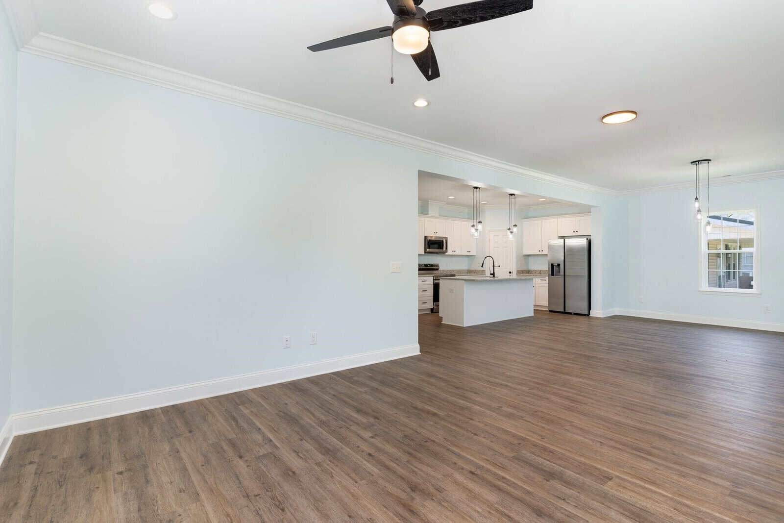 Spacious open-concept living area with light wood flooring and soft blue walls. Modern kitchen in the background featuring white cabinets, stainless steel appliances, and an island. Ceiling fan and recessed lighting adorn the room.