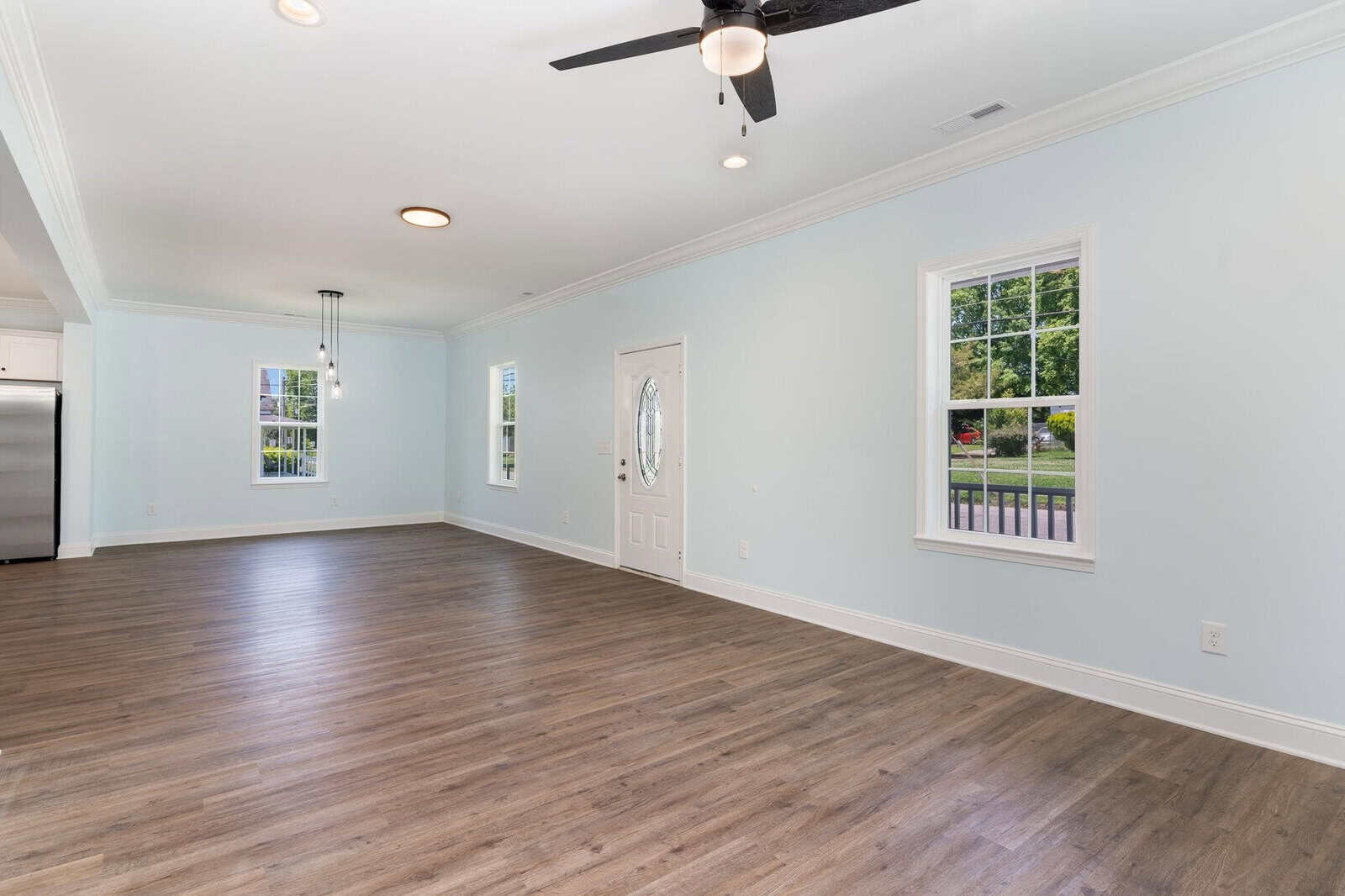 Spacious empty room with light blue walls, wood flooring, and large windows. Features a ceiling fan with lights, a modern pendant light fixture, and a white front door with an oval glass insert. Natural light fills the room.
