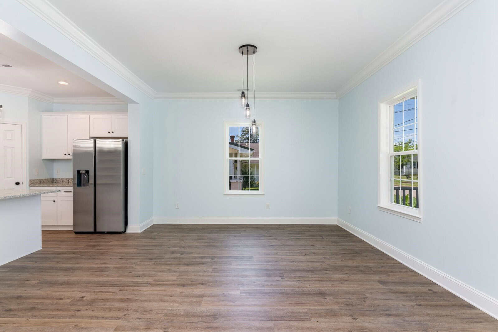 A modern, unfurnished living room with light blue walls, wooden floors, and a decorative ceiling light. Large windows provide natural light. The open kitchen area features white cabinets and a stainless steel refrigerator.