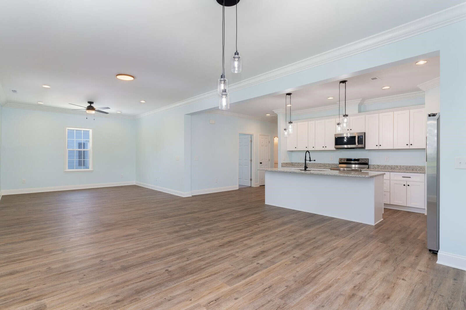Spacious open-concept room with light hardwood floors, pale blue walls, and recessed lighting. Features a modern kitchen with white cabinets, granite countertops, and pendant lights. A ceiling fan is centered above the living area.