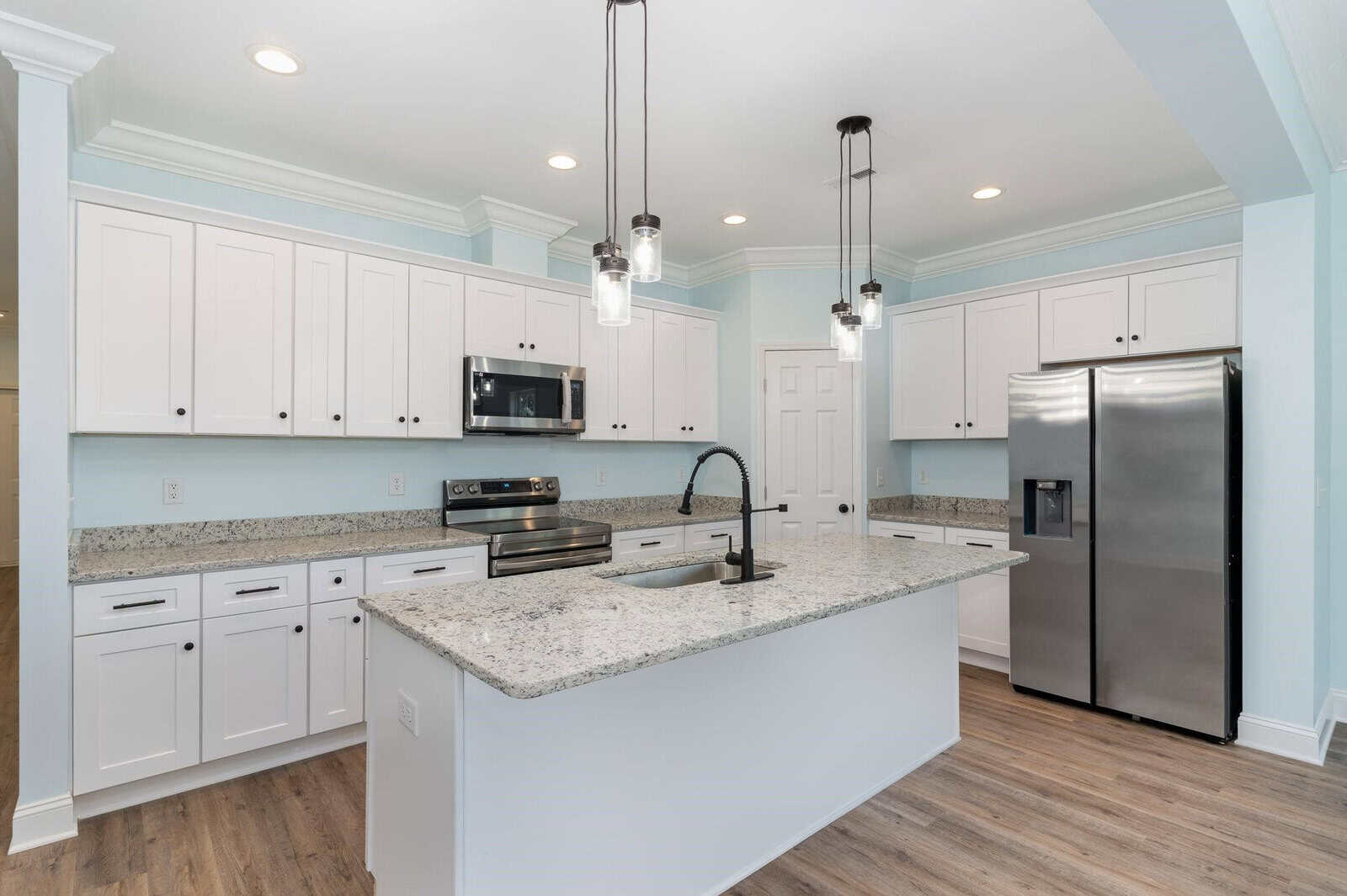 Modern kitchen with white cabinets, stainless steel appliances, and a granite island. Pendant lights hang above the island. Light blue walls and wood flooring complete the space.