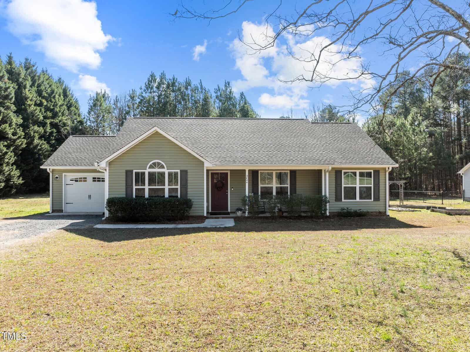 A single-story green house with white trim graces 3044 Cannady Mill Road in Franklinton. Nestled in front of a wooded area, it features a garage on the left, a welcoming front porch with a red door, and a large window. The yard is grassy under a clear sky dotted with a few clouds.