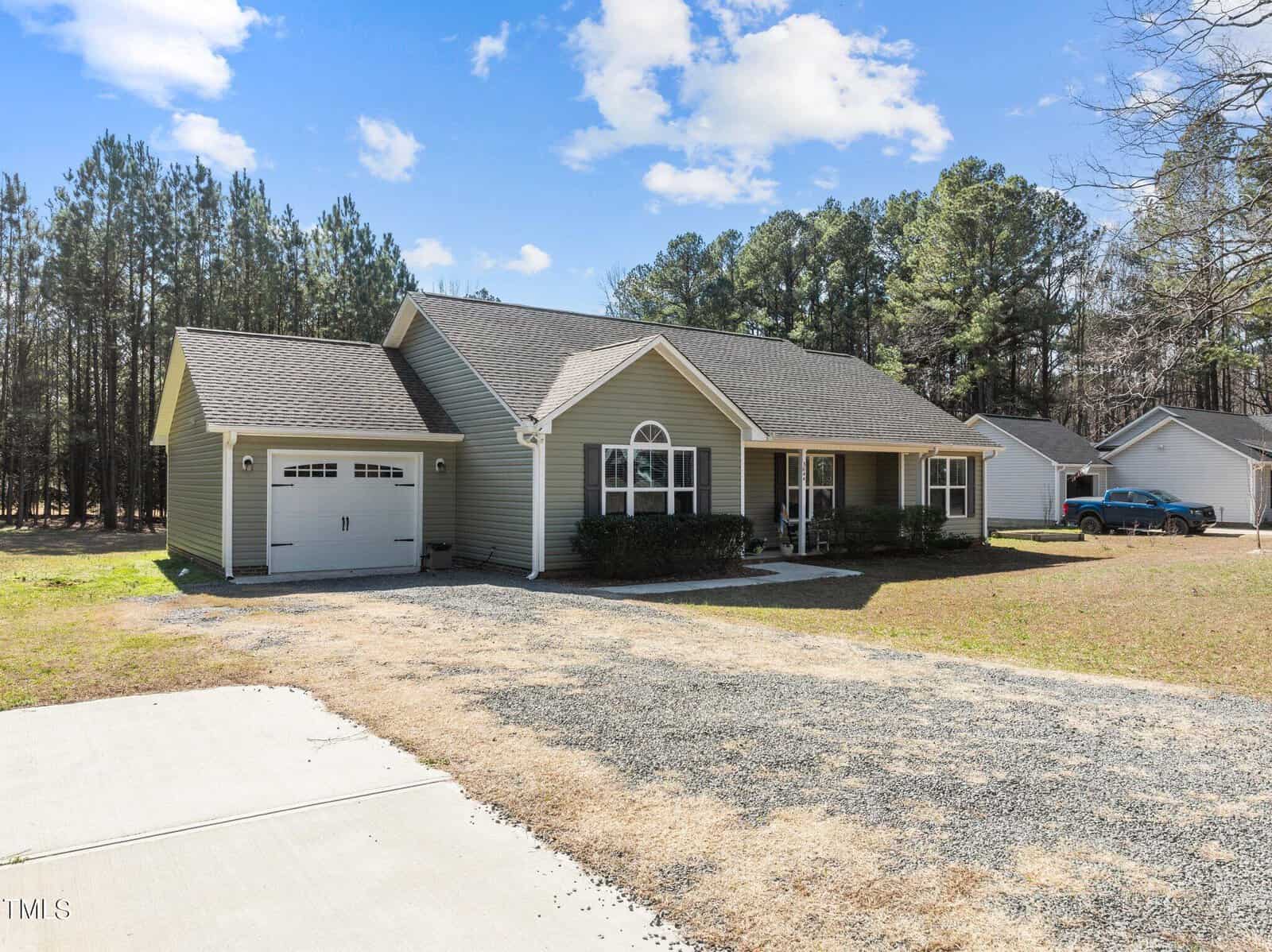 A single-story house with a gray exterior and attached garage sits in a grassy yard bordered by a wooded area. A paved driveway leads to the garage. The sky is partly cloudy, and there is a parked car in the background.